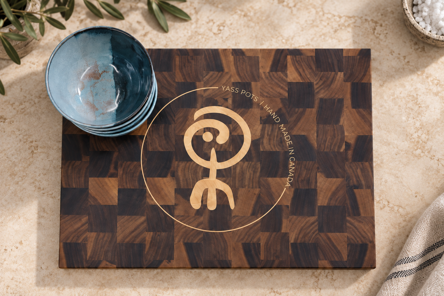 Wooden cutting board with a logo, stack of blue ceramic bowls, and a bowl of white pebbles, on a beige surface with a cloth and green plants nearby.