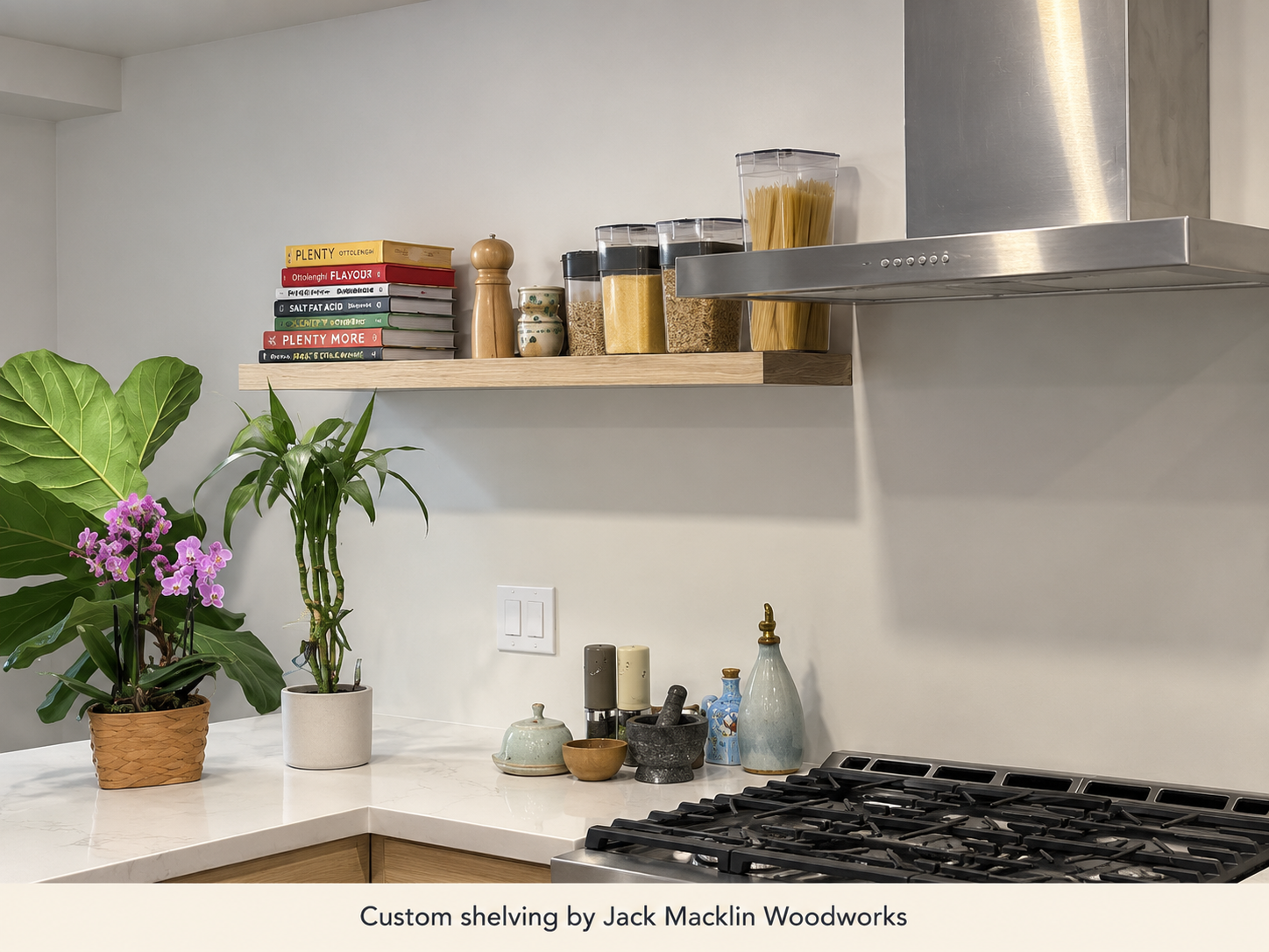 A kitchen countertop with potted plants, spice jars, and kitchen tools. Above the countertop, a wooden shelf holds cookbooks, jars of pasta and grains, and a pepper grinder. A stainless steel range hood is visible on the right.