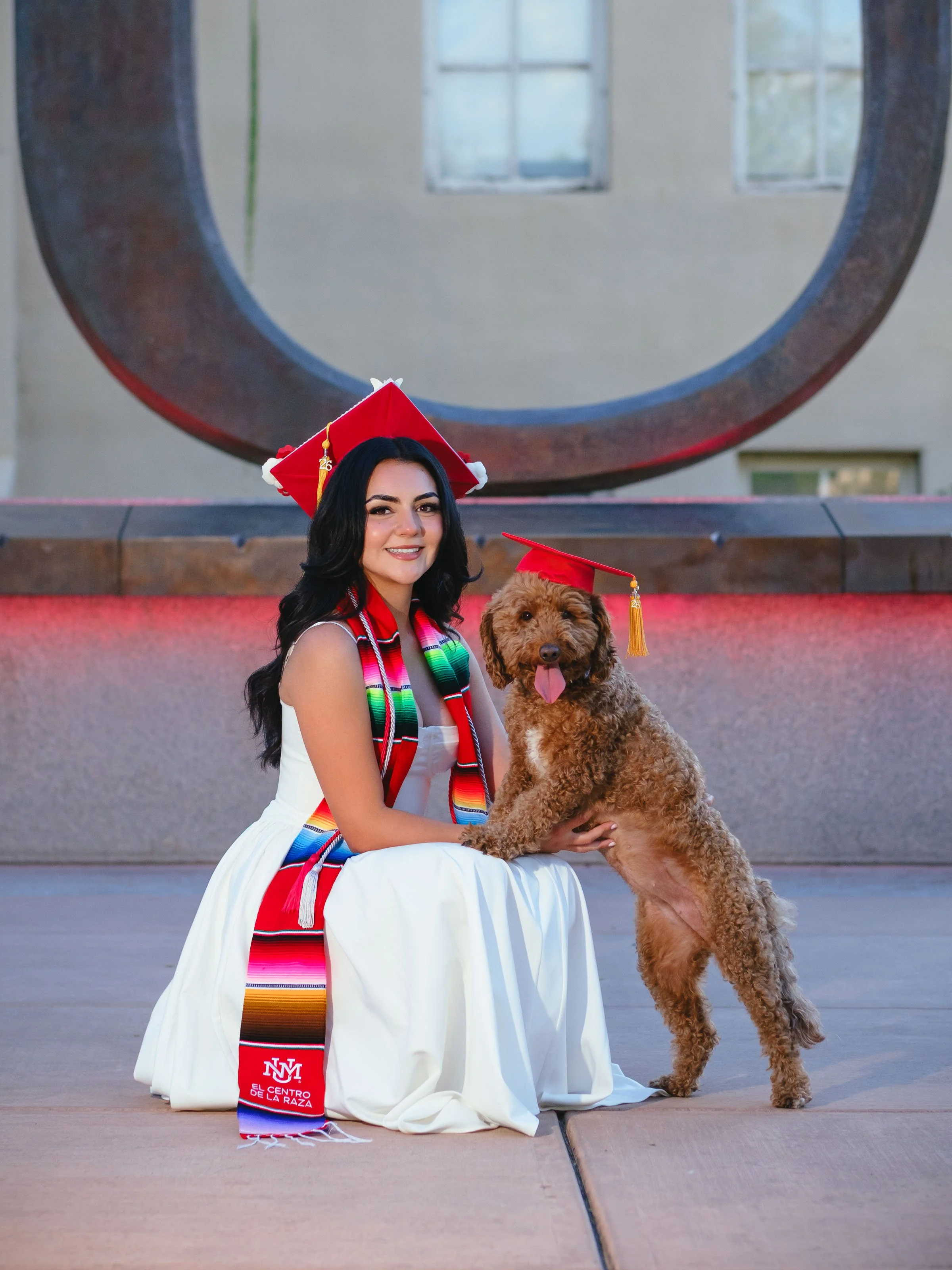 University of New Mexico graduate wearing a Centro de la Raza stole seated with her dog in front of the Big U during a UNM graduation portrait session celebrating the Raza Stoling Ceremony in Albuquerque.