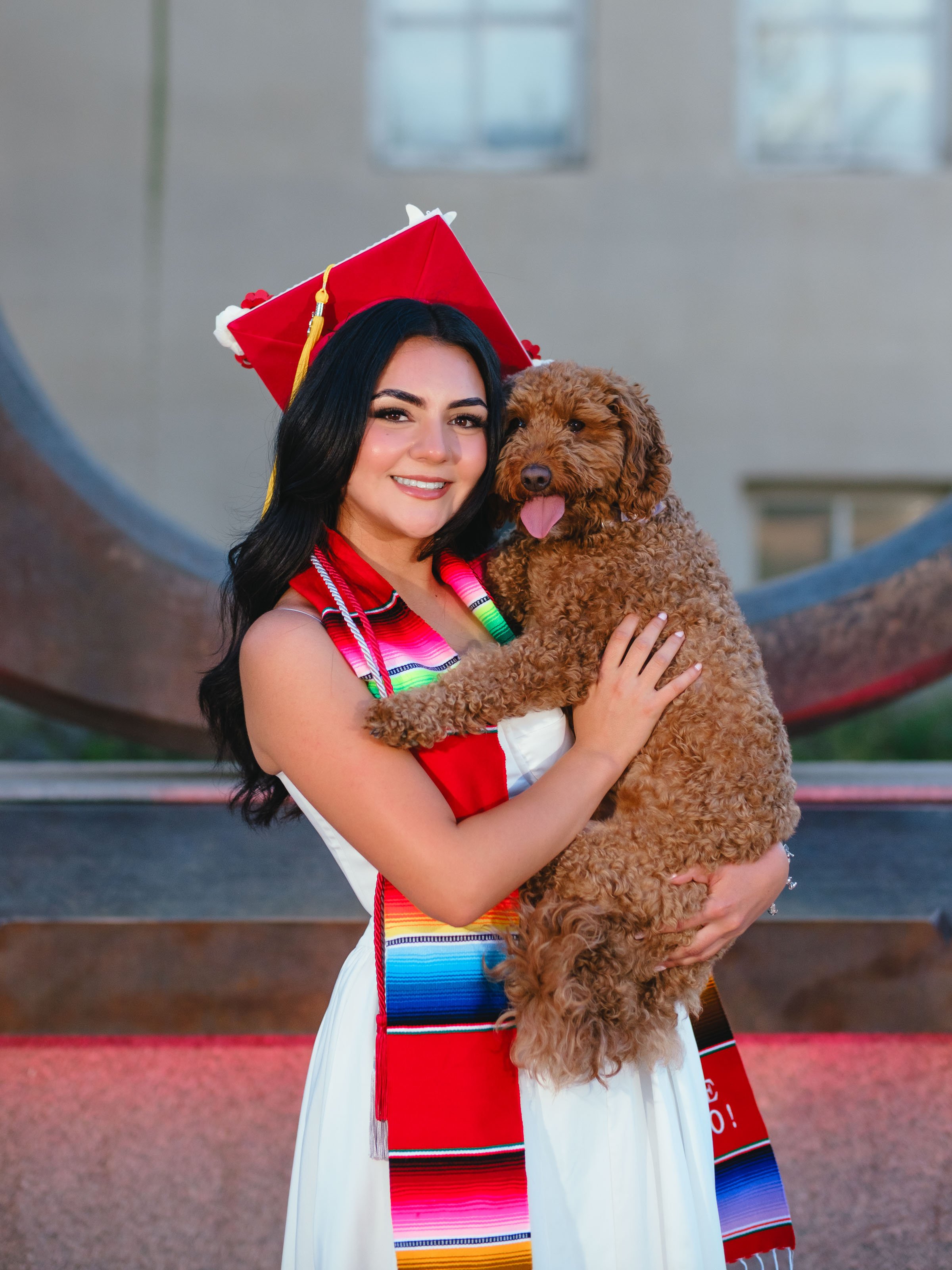 UNM graduate wearing a Centro de la Raza stole posing with her dog in front of the Big U during a University of New Mexico Raza Stoling Ceremony graduation photo session in Albuquerque.
