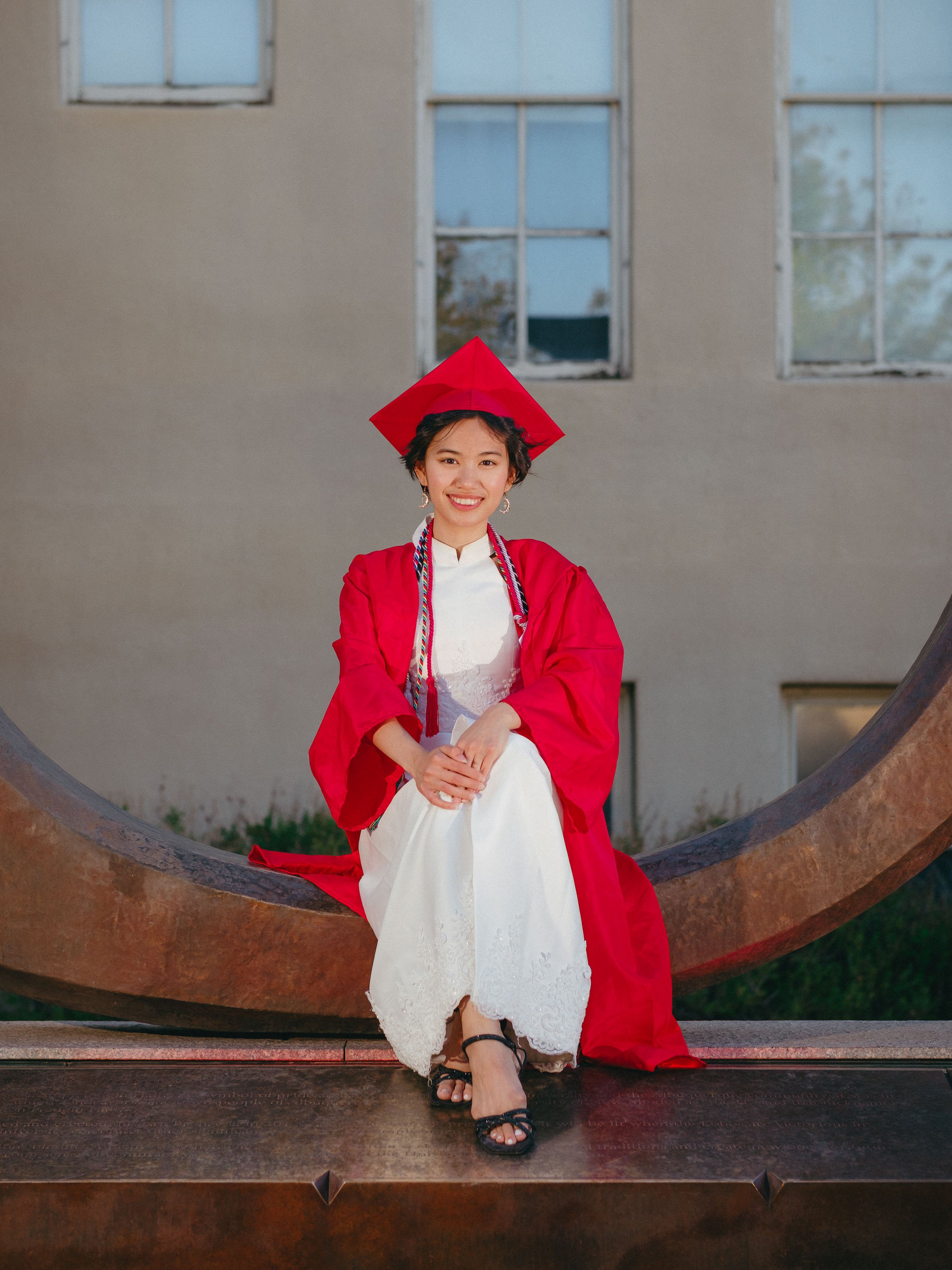 UNM graduate seated in front of the Big U wearing a red cap and gown during a University of New Mexico graduation portrait session in Albuquerque.