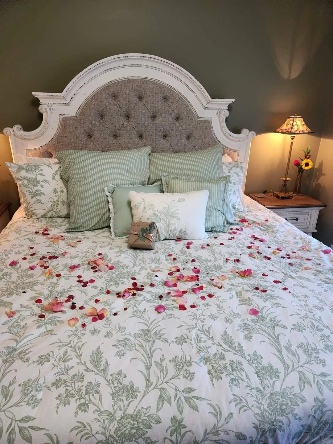 A bed in the cottage with a floral sheet, rose petals, and multiple pillows, featuring a white and beige headboard; side table with a lamp and small flower arrangement.
