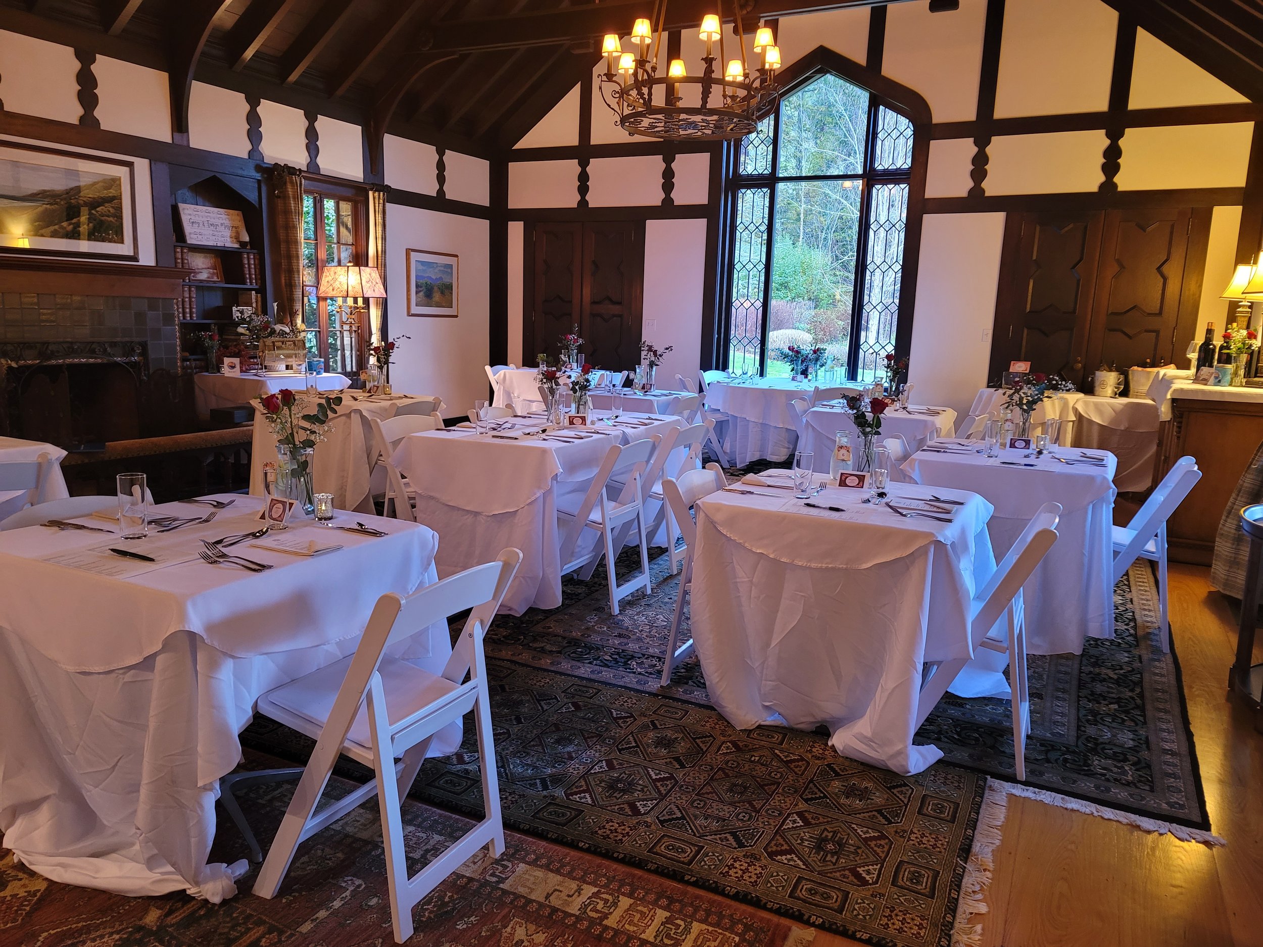 A room decorated for an event with several round tables covered with white tablecloths, each set with silverware, glasses, and a small floral centerpiece. The room has high, dark wood-paneled walls, large windows, and a decorative chandelier hanging from a vaulted ceiling. There is a fireplace with a framed picture above it and a side table with bottles of wine and other items.