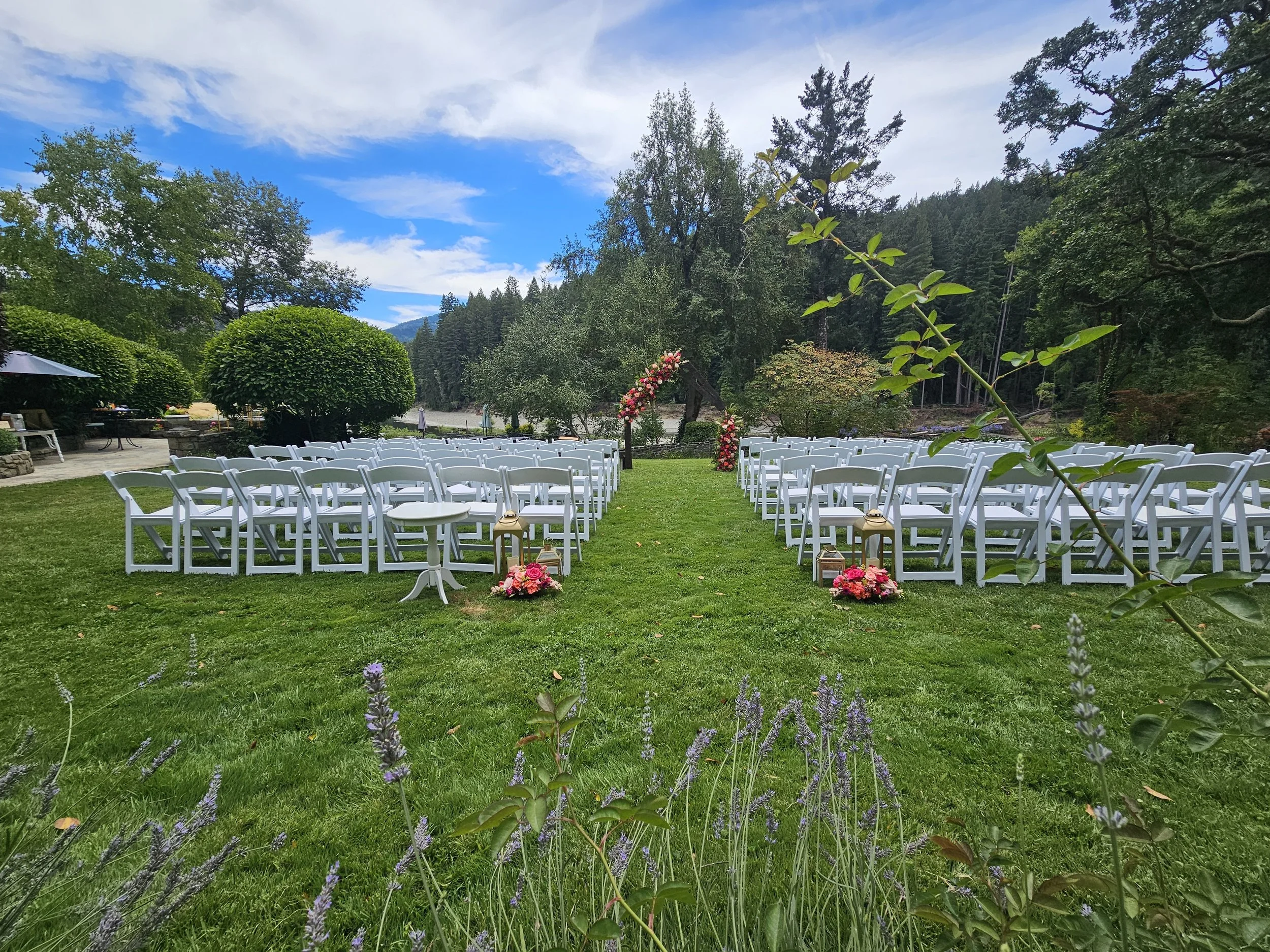 Outdoor wedding setup with white chairs arranged on green grass, decorated with pink flowers, in a scenic setting with trees, flowers, and mountains under a partly cloudy sky.