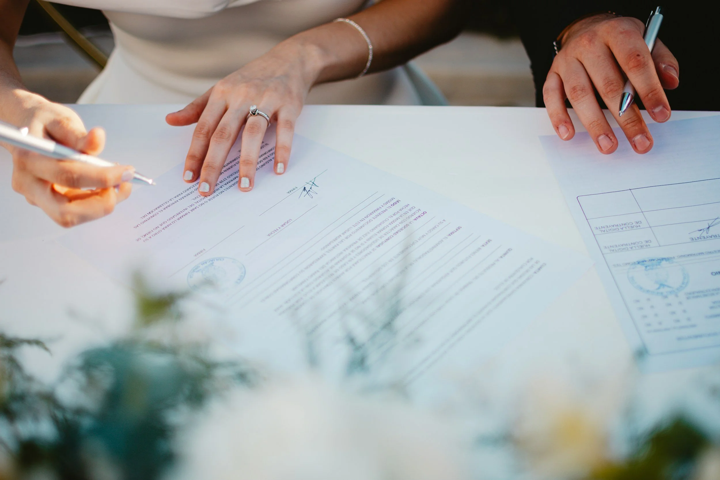 Bride and groom sitting at a white table.   There is a pen in her left hand and a pen in his right hand and they are both signing a marriage license.