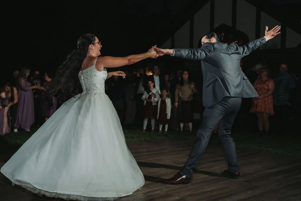 Couple dancing at a wedding reception, with the bride in a white wedding gown and the groom in a gray suit, holding hands and dancing on a wooden floor, surrounded by guests.