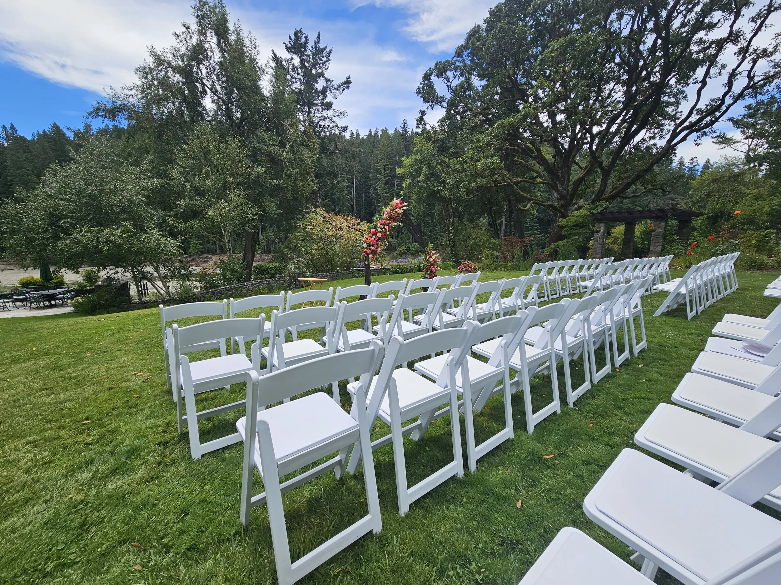 Outdoor wedding setup with rows of white chairs on a grassy lawn, decorated with flowers, in front of large trees and a forested background under a cloudy sky.