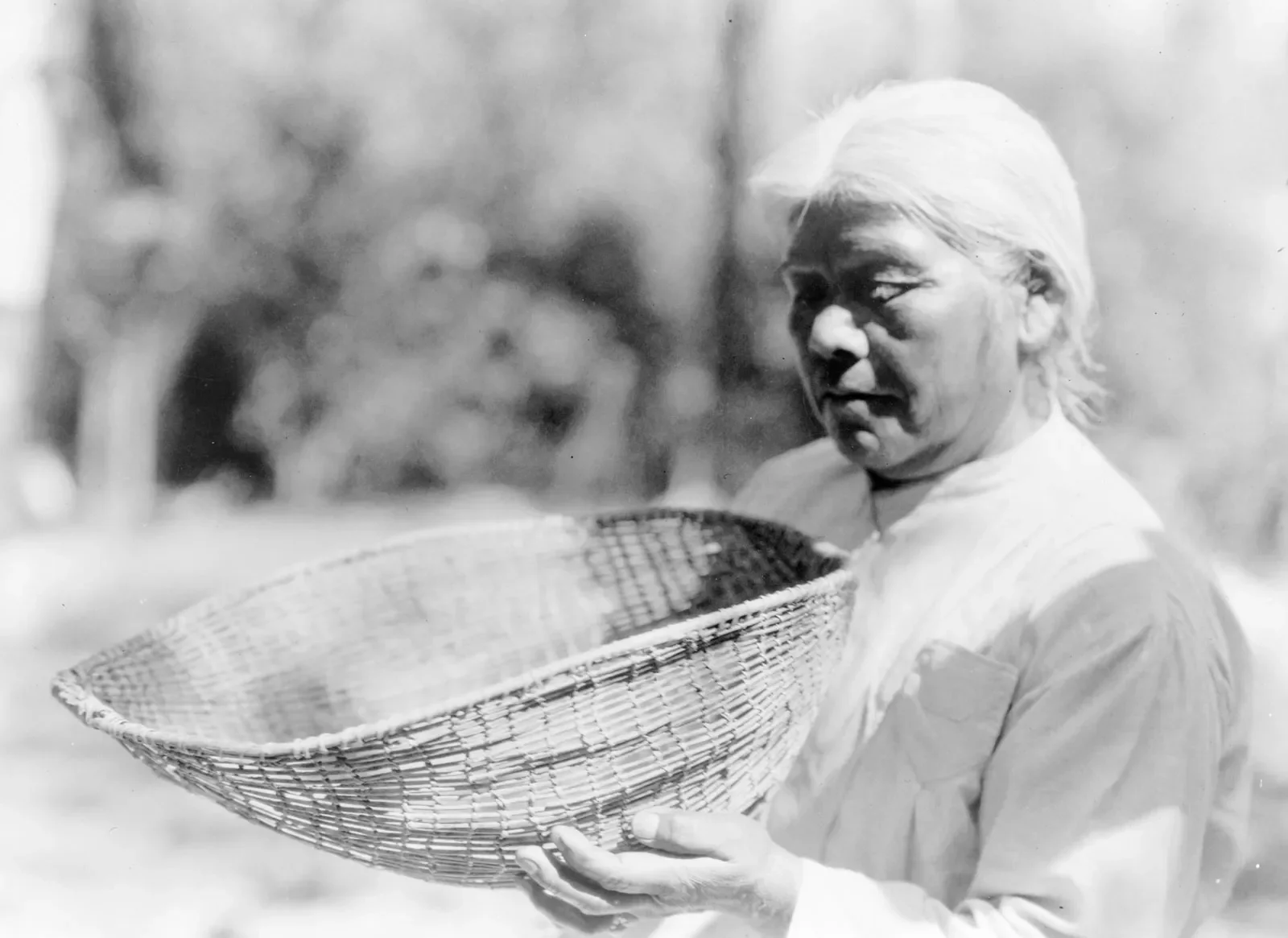 An elderly indigenous woman with white hair holds a woven basket and looks downward with a serious expression, outdoors with blurred trees in the background.