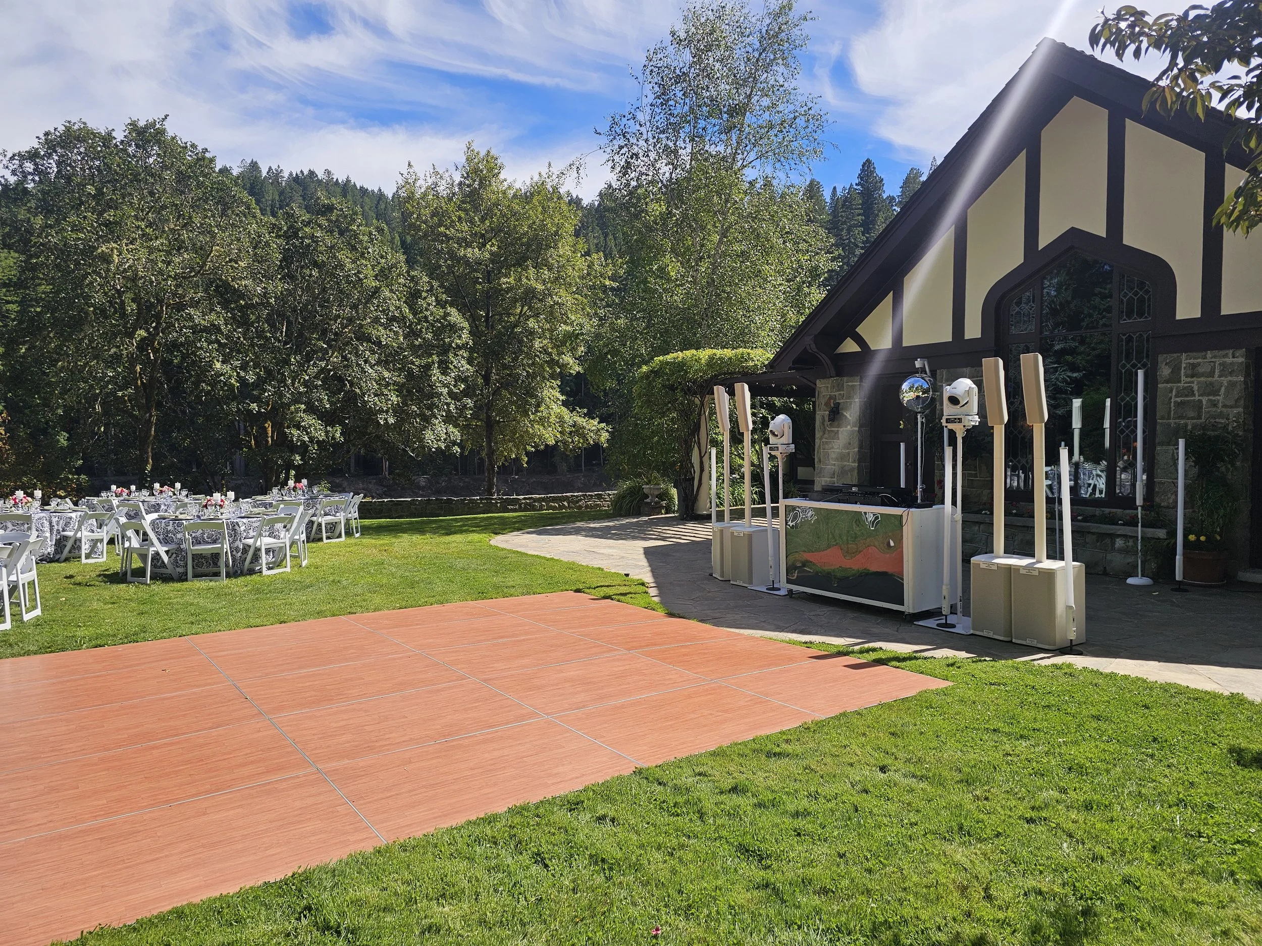 An outdoor event setup with round tables covered in blue patterned tablecloths and white chairs, set on a grassy lawn with a backdrop of tall trees and a partly cloudy sky. A building with dark brown beams and light walls is visible to the right, along with audio equipment and speakers indicating preparations for a celebration or gathering.