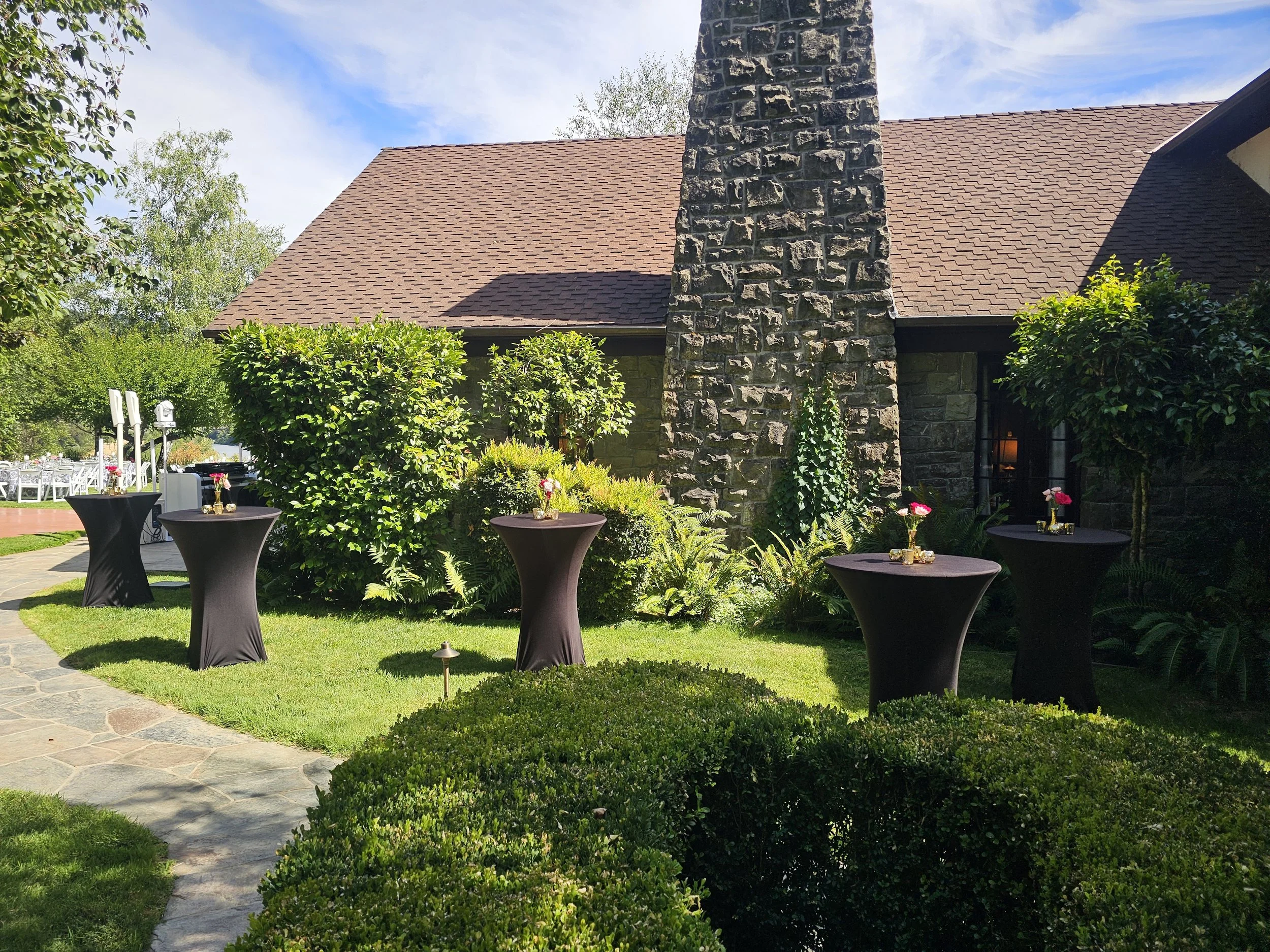 Outdoor garden scene with manicured bushes, round tables covered with black tablecloths and gold vases with pink flowers, stone pathway, trees, house with stone chimney and brown shingle roof, and a clear partly cloudy sky.