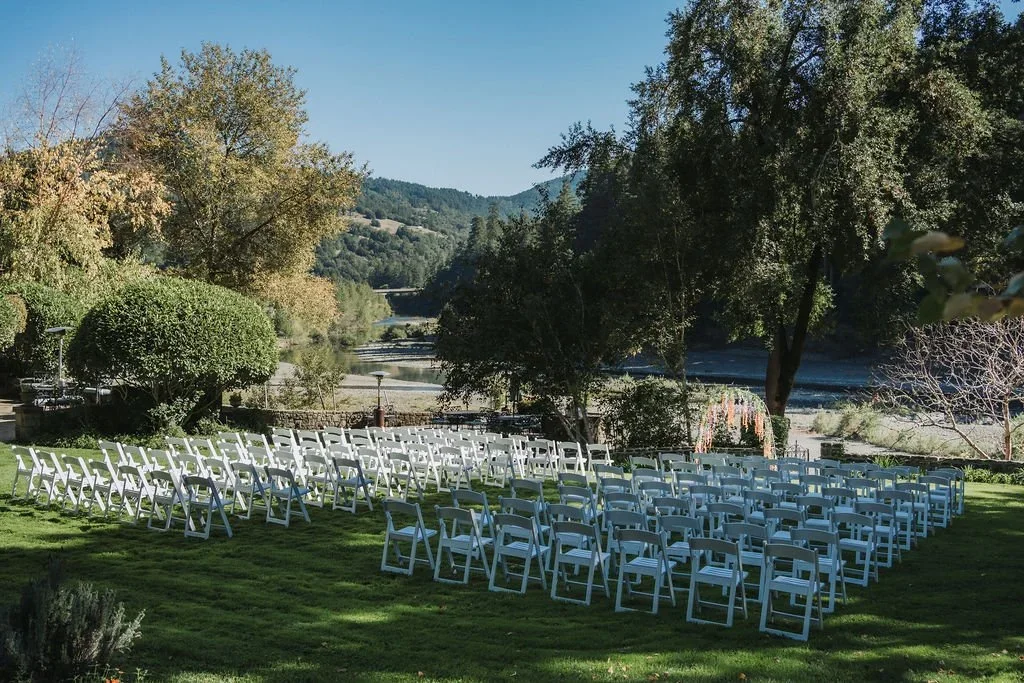 Outdoor wedding ceremony space with rows of white chairs on a grassy hill overlooking a river, surrounded by trees and hills.