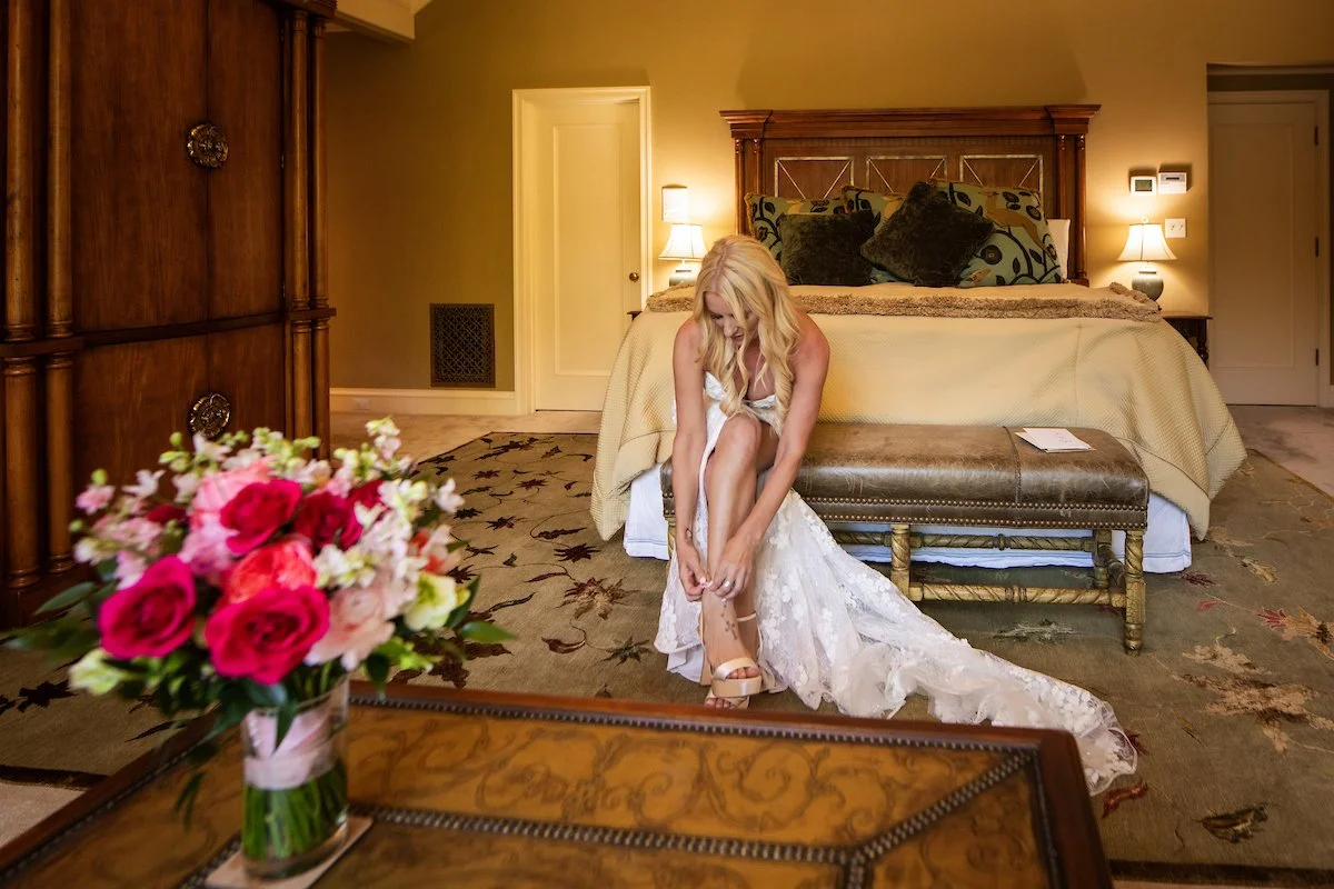 A woman in a wedding dress sits on a bench in a hotel bedroom, adjusting her shoe, with a bouquet of pink and white flowers on a table in the foreground. Photo by Moonstone images.