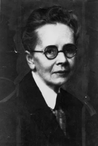 A black-and-white photo of a woman, Julia Morgan, wearing glasses, with short, wavy hair, dressed in a dark coat and white shirt.
