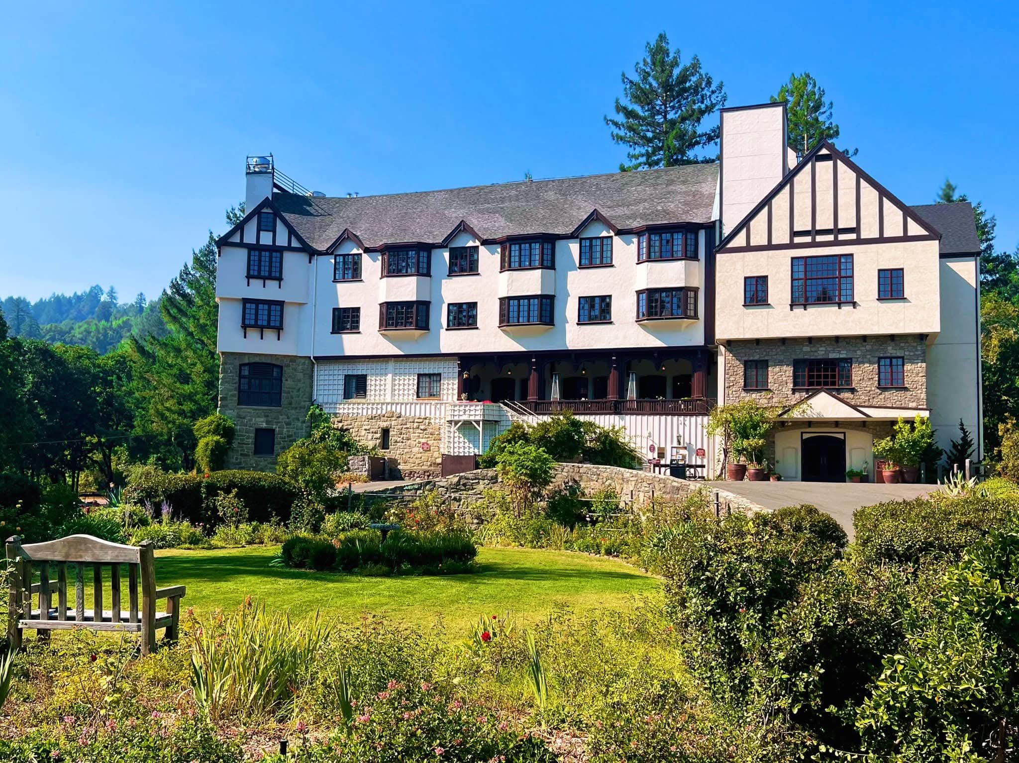 Benbow Historic Inn, big historic building with white walls, dark wood trim, multiple bay windows, and a stone lower level, surrounded by lush garden and green trees under a clear blue sky.