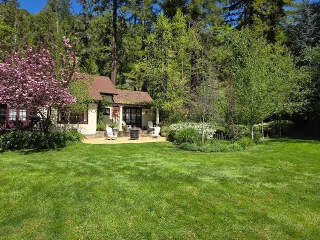 A backyard with a well-maintained lawn, a patio area with outdoor chairs, a small grill, and a house in the background surrounded by trees and blooming flowers.