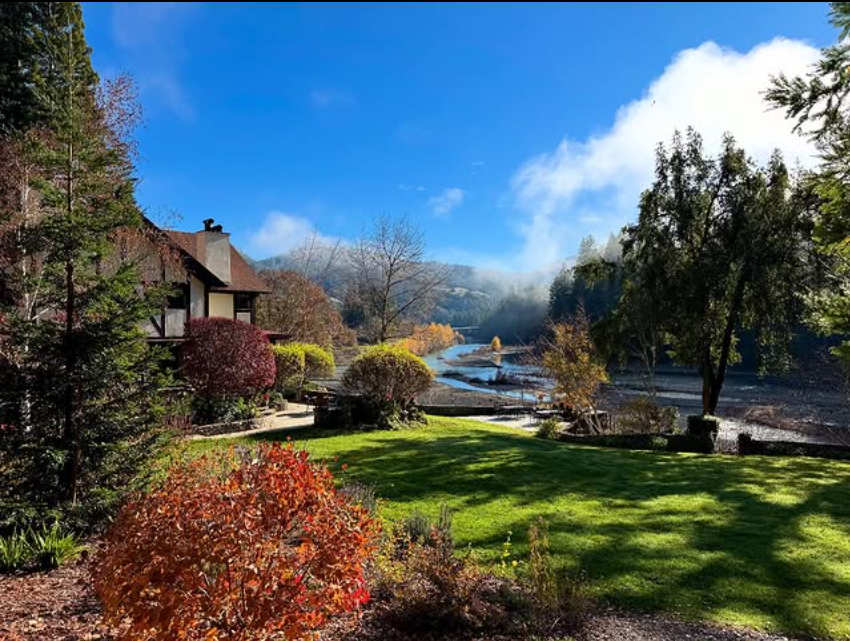 A scenic view of a lush garden at the historic Julia Morgan Redwood Grove estate with colorful bushes and trees, the Julia Morgan Redwood Grove house on the left, the Eel river flowing in the background, and a bright blue sky with some clouds.