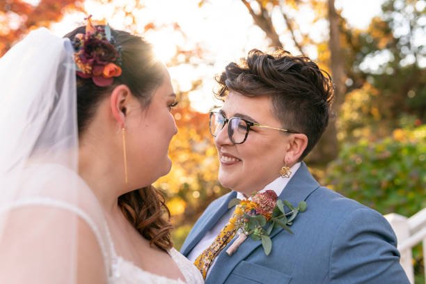 Two women, one in a wedding dress and the other in a blue suit, smiling at each other outdoors at the Julia Morgan Redwood Grove property.