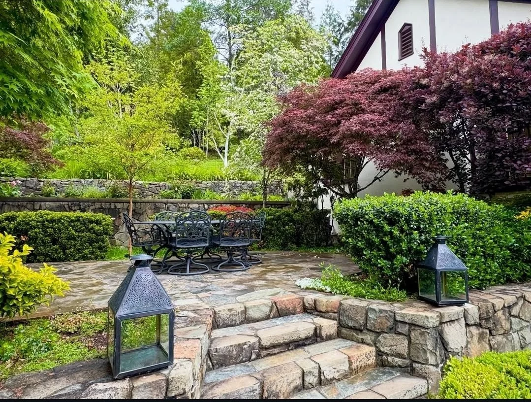 A lush backyard garden terrace with stone steps, black metal outdoor furniture, two lanterns, and a variety of green and purple bushes and trees.