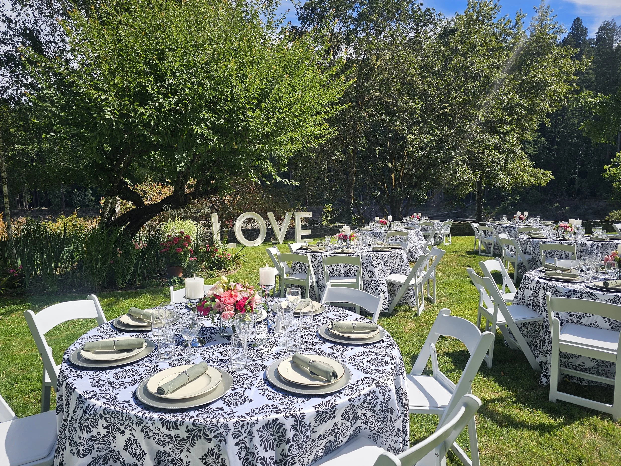 Outdoor wedding reception setup with round tables covered in black and white patterned tablecloths, white chairs, and floral centerpieces, set in a lush green garden with trees and a blue sky.