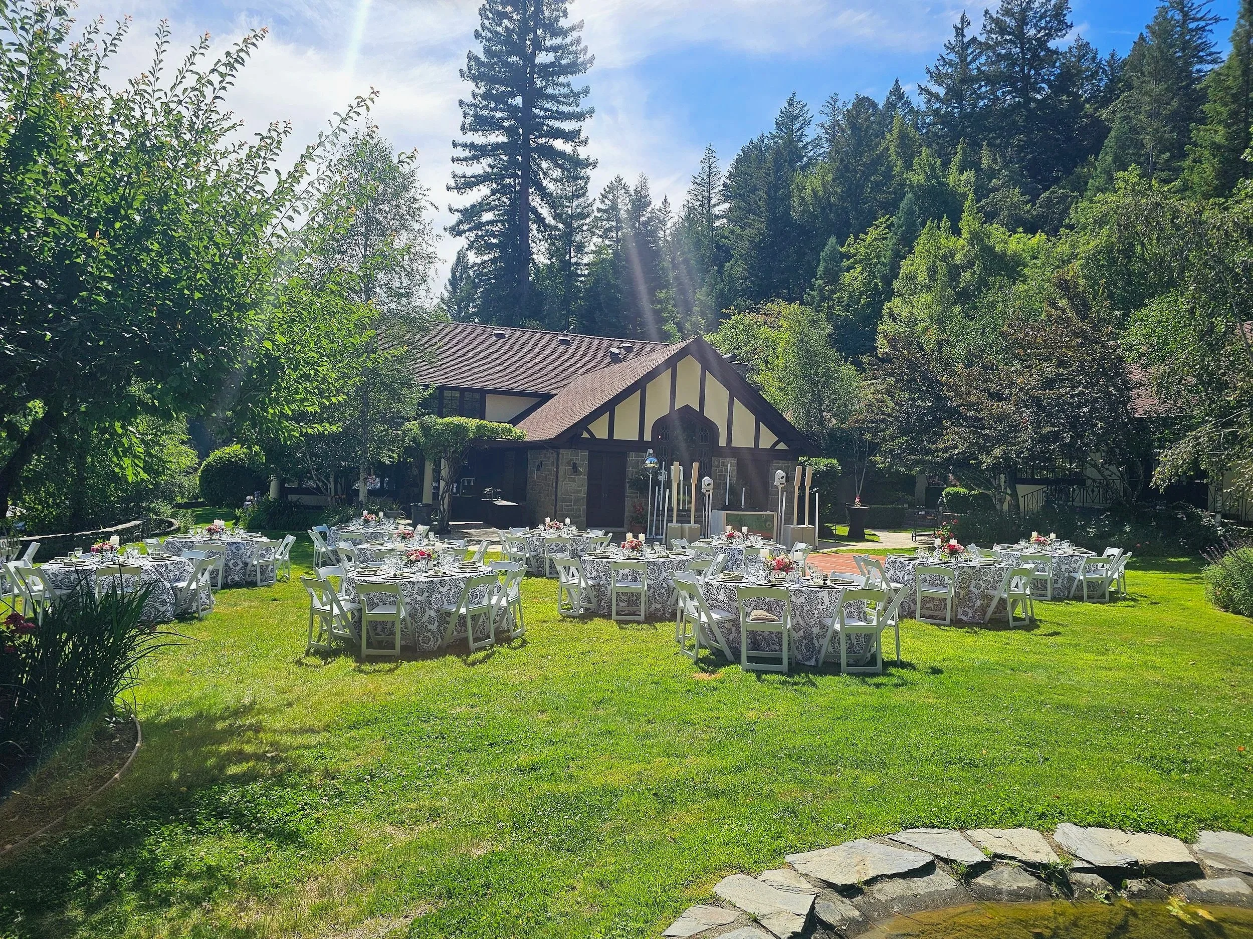 An outdoor event setup in a lush green garden with white tables and chairs decorated with pink flowers, surrounded by tall trees and a house in the background under a blue sky.