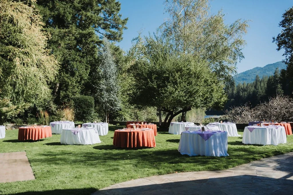 Outdoor event setup with round tables draped in white and orange tablecloths, nestled on a grassy lawn surrounded by trees, under a clear blue sky.