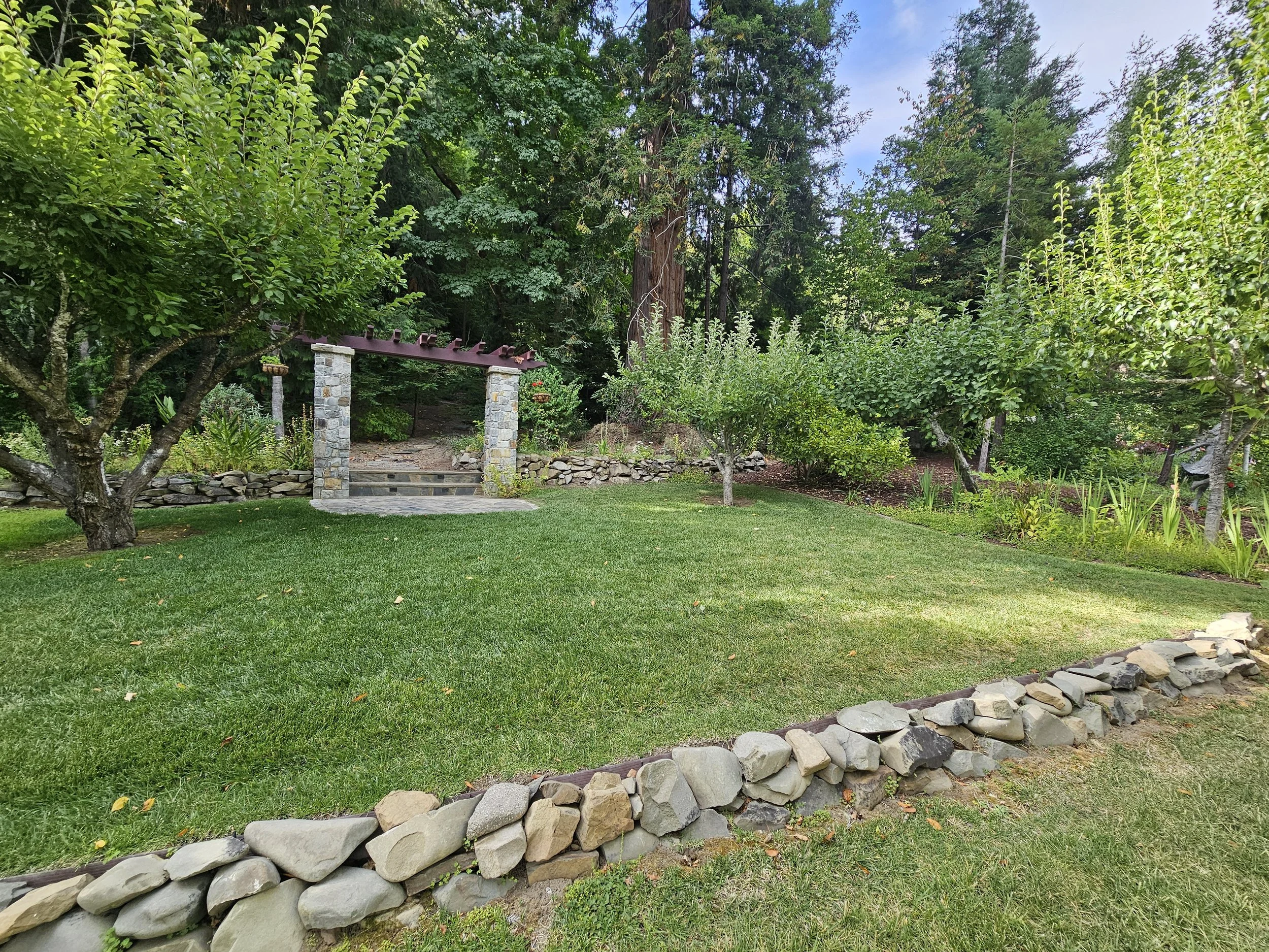 A small orchard area with a green lawn, multiple trees, a stone pathway, and a wooden arbor supported by stone pillars, with dense forest in the background.