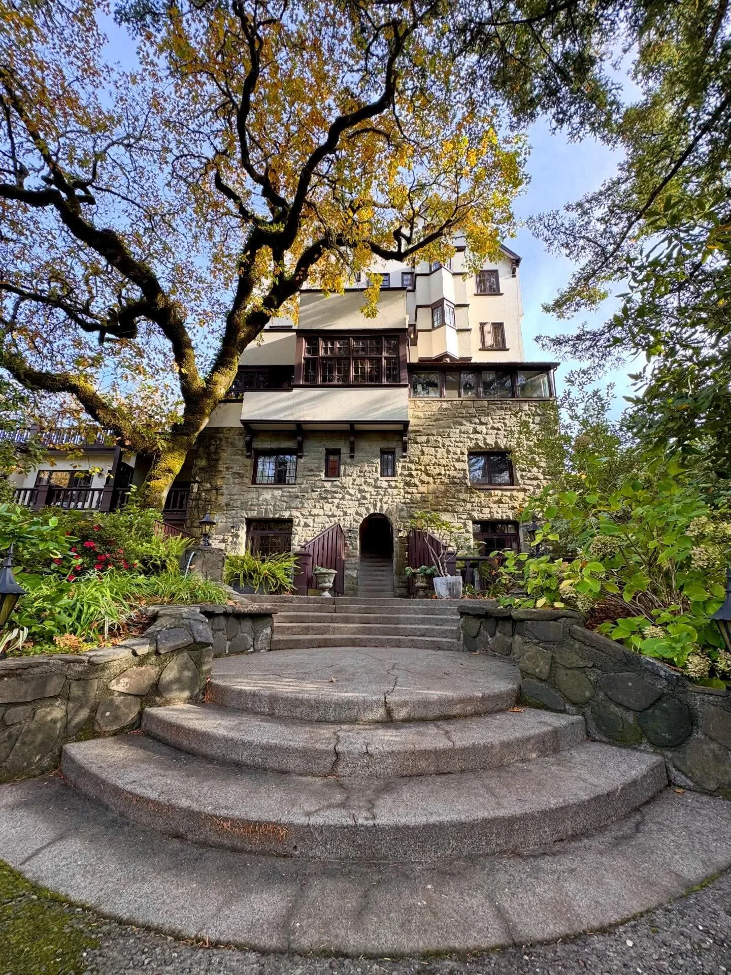 A large multi-story Benbow Historic Inn, with a stone and white exterior, surrounded by trees and landscaped garden, with steps leading up to the entrance.