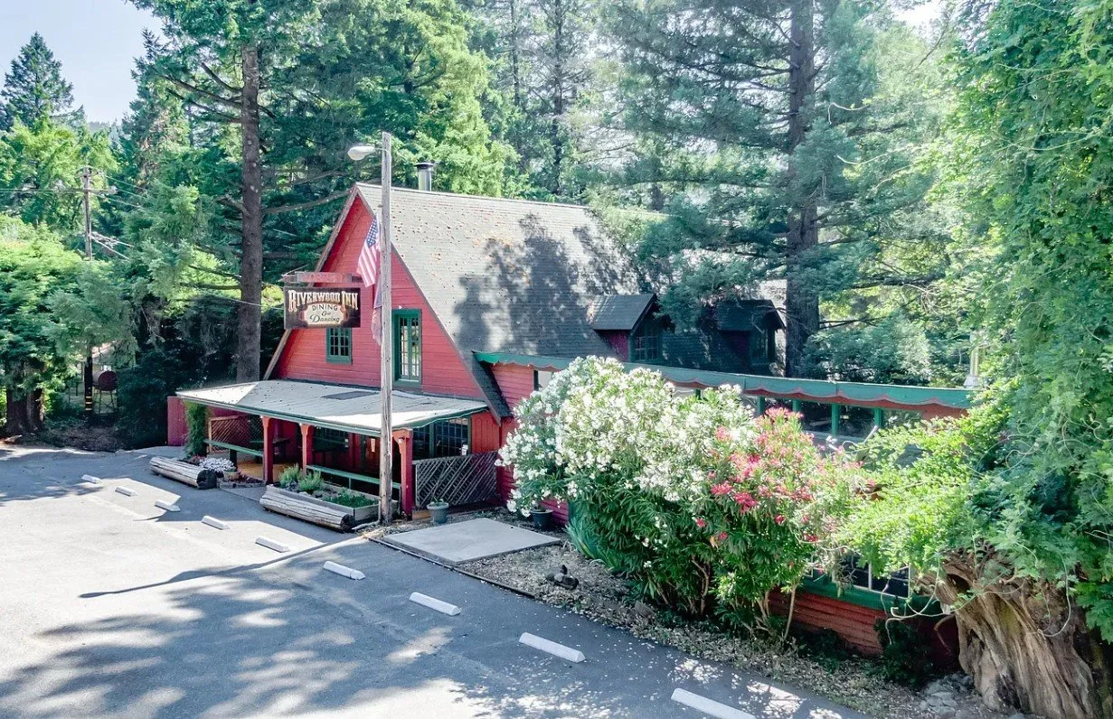 A red wooden building labeled Riverwood Inn with a covered porch, surrounded by tall green trees and flowering bushes, next to a parking lot.