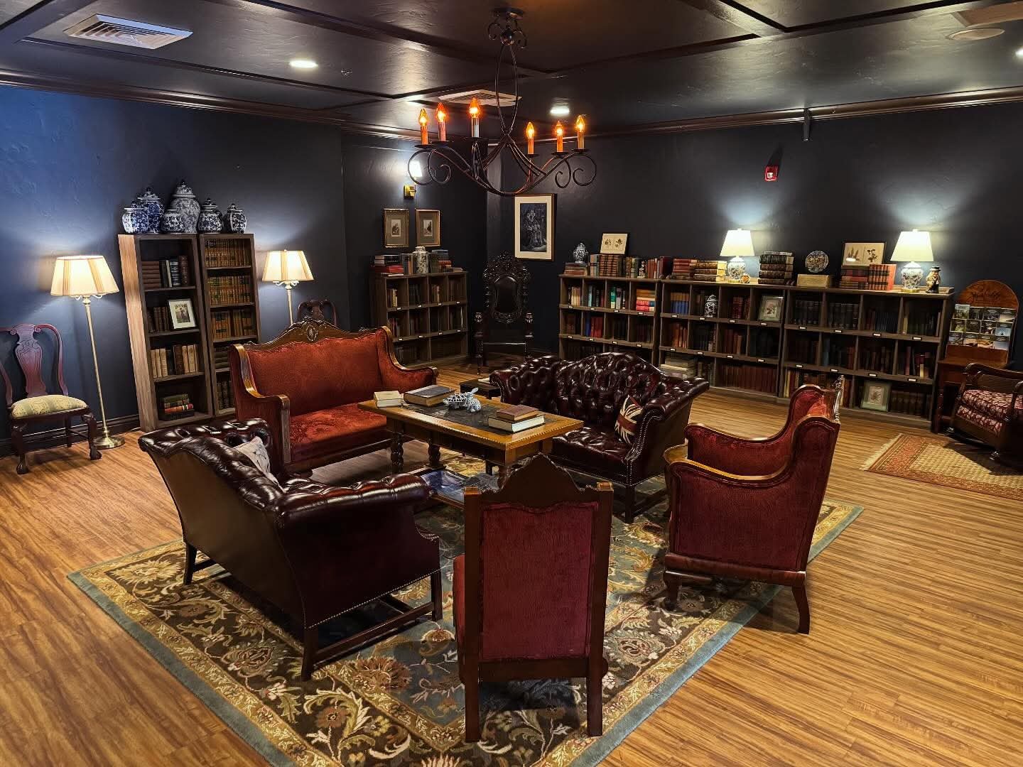 A cozy, vintage-style library room with dark walls, wooden flooring, and classic furniture. Central area features a round table surrounded by leather and upholstered armchairs, with books and decorative items on top. Bookshelves filled with books line the back wall, illuminated by table lamps and a chandelier hanging from the ceiling.