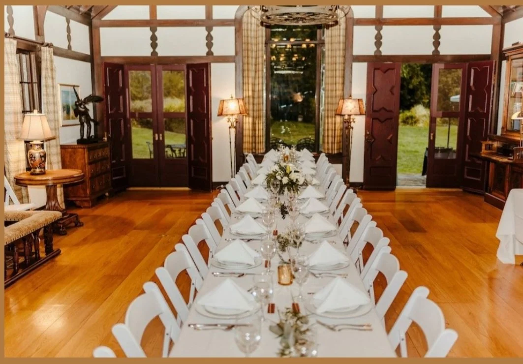 A long dining table set with white napkins, glassware, and floral centerpieces in a historic great room with wooden floors, large windows, and warm lighting.