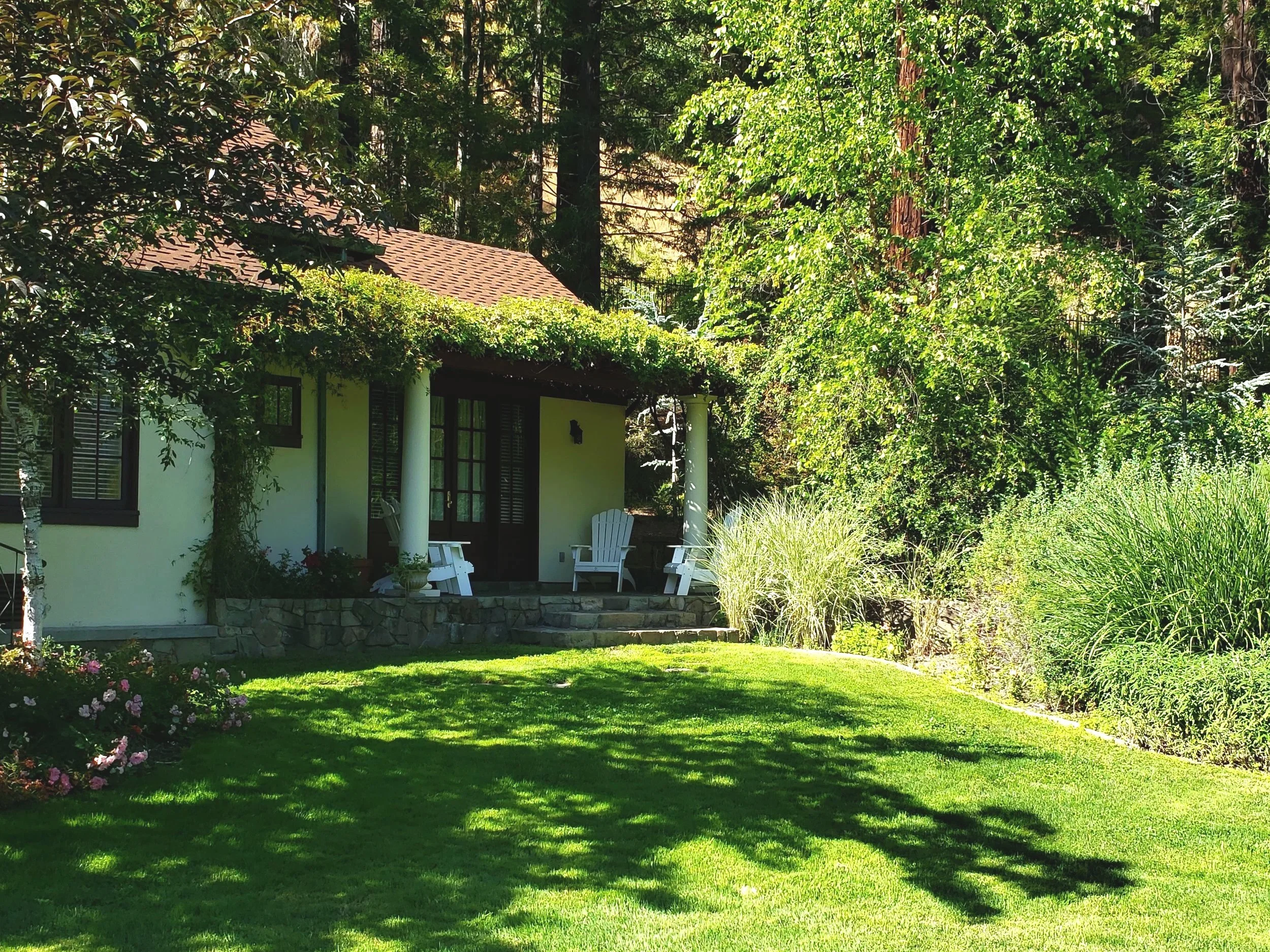 A house with a light yellow exterior, dark brown roof, white columns, and surrounded by lush green trees and well-maintained lawn with sunny weather.
