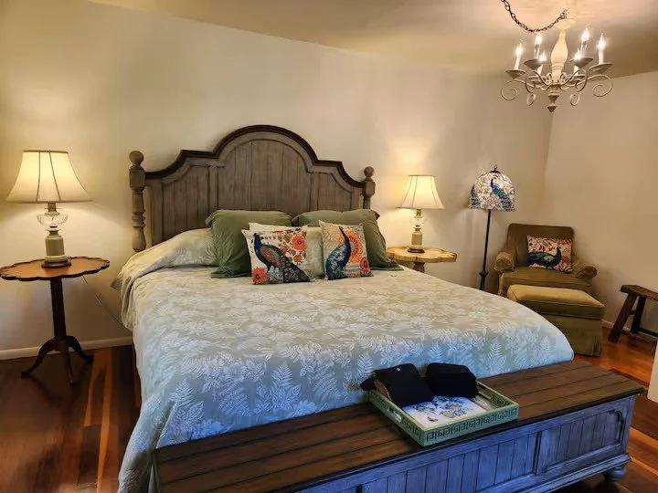 A bedroom with a wooden bed, two nightstands with lamps, a green armchair, a colorful lamp, and a chandelier.