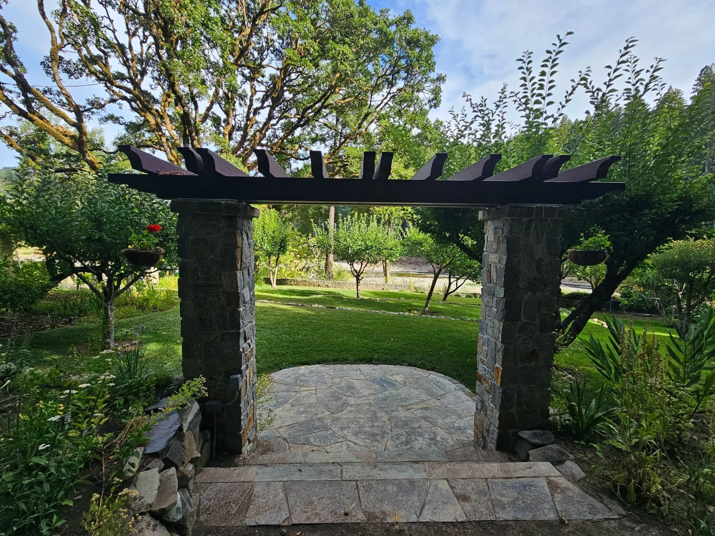 Stone archway with a wooden top, surrounded by green trees and plants, with a garden and pathway beyond.
