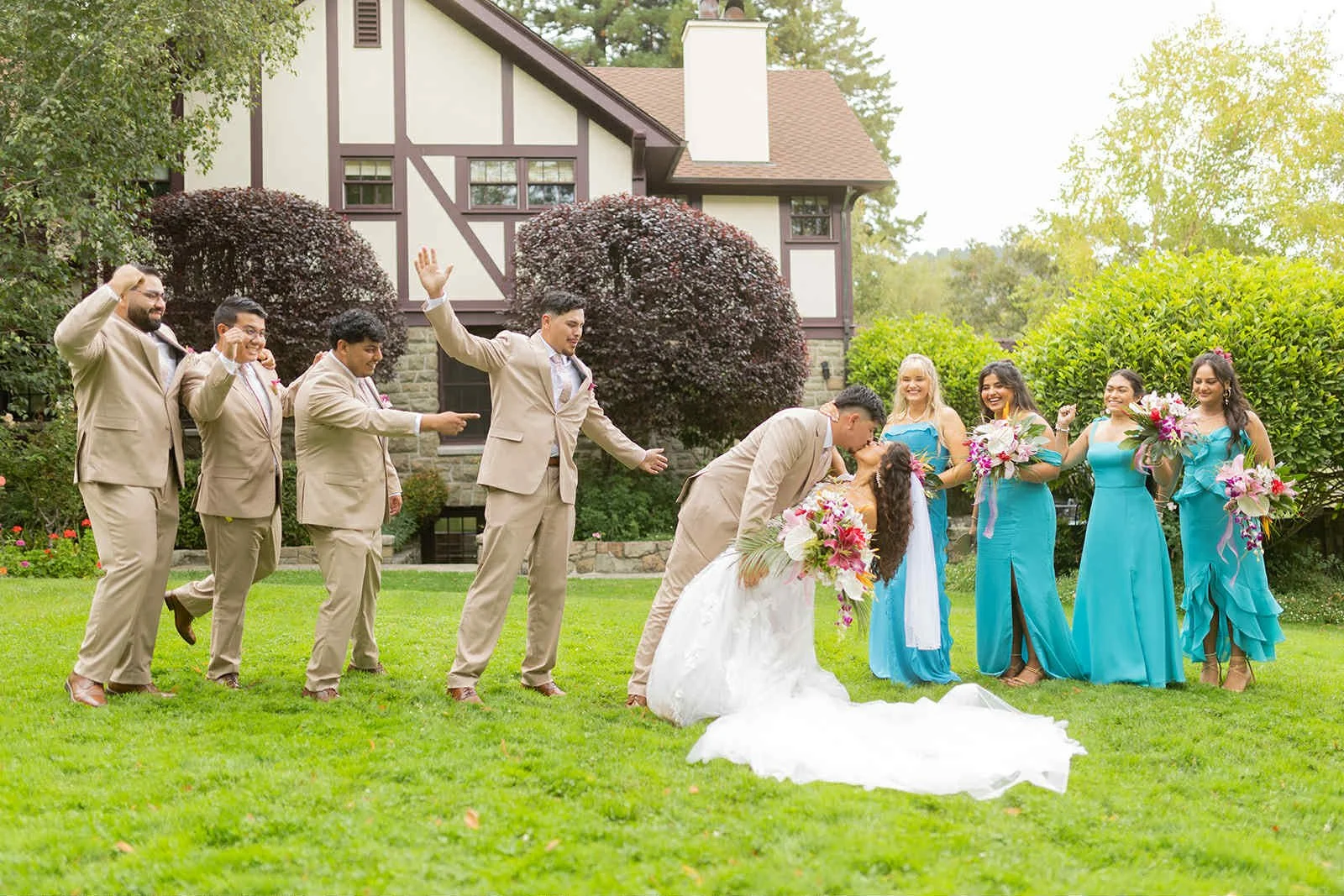 A bride and groom kissing outdoors on their wedding day, surrounded by bridesmaids in turquoise dresses and groomsmen in beige suits, with a house and lush greenery in the background.