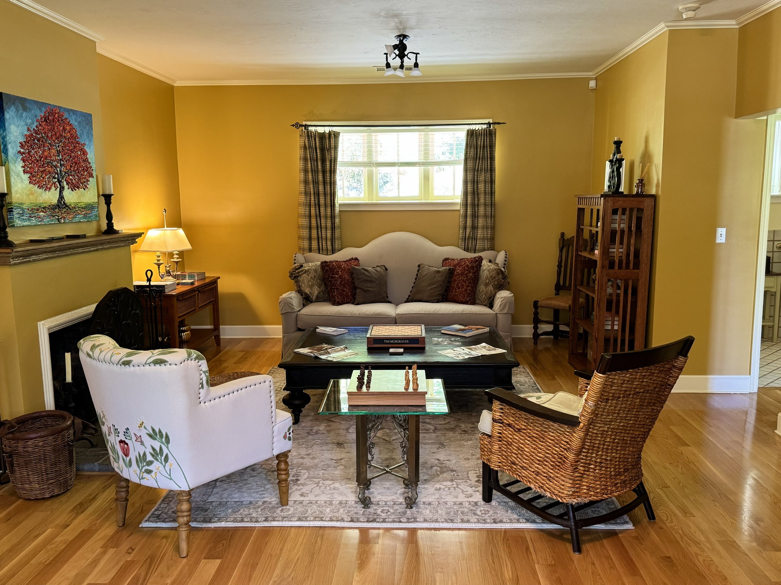 Living room with yellow walls, wooden floor, and various chairs including a white armchair with floral upholstery, a wicker armchair, and a black chair. There is a glass-top coffee table with books and a chess set, a sofa with decorative pillows, a window with plaid curtains, and artwork on the walls. A ceiling light fixture is visible.