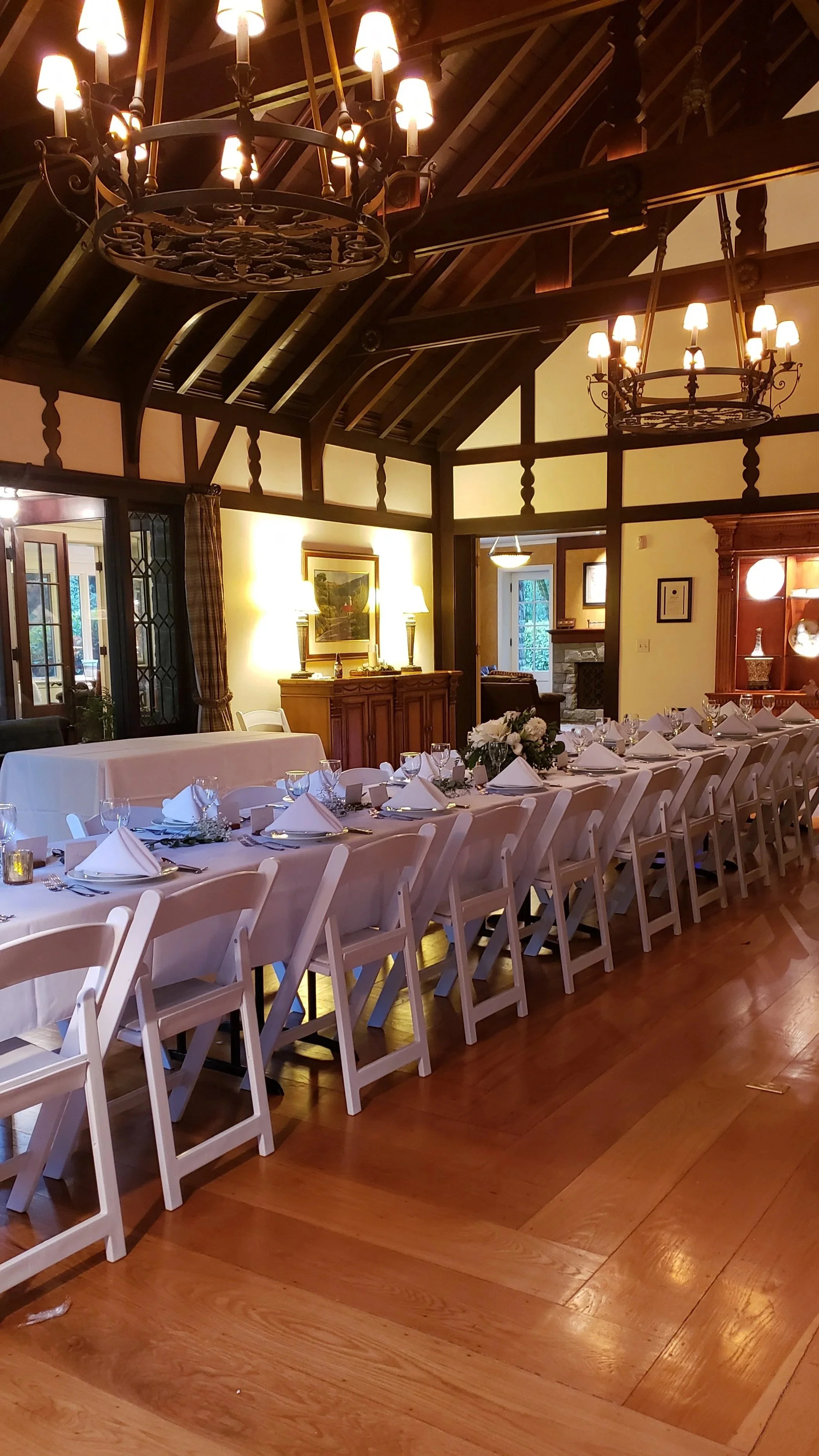 A great room with a long table set for a wedding reception meal, decorated with white tablecloths, napkins, glassware, and floral centerpieces. The room has wooden walls, chandeliers hanging from the ceiling, and a fireplace in the background.