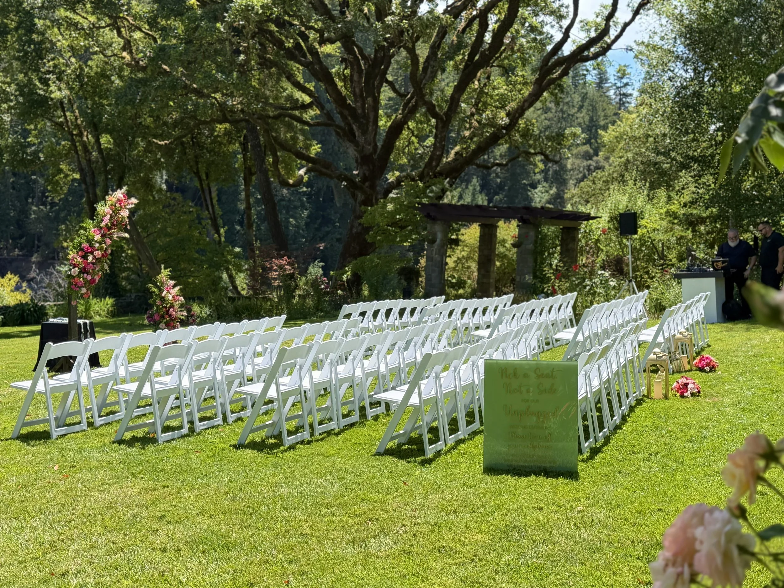 An outdoor wedding ceremony setup with white folding chairs arranged on a grassy lawn, decorated with pink and white floral arrangements. There is a large mature tree providing shade and a DJ booth with two people setting up in the background. A green sign with wedding-related text is placed at the front.