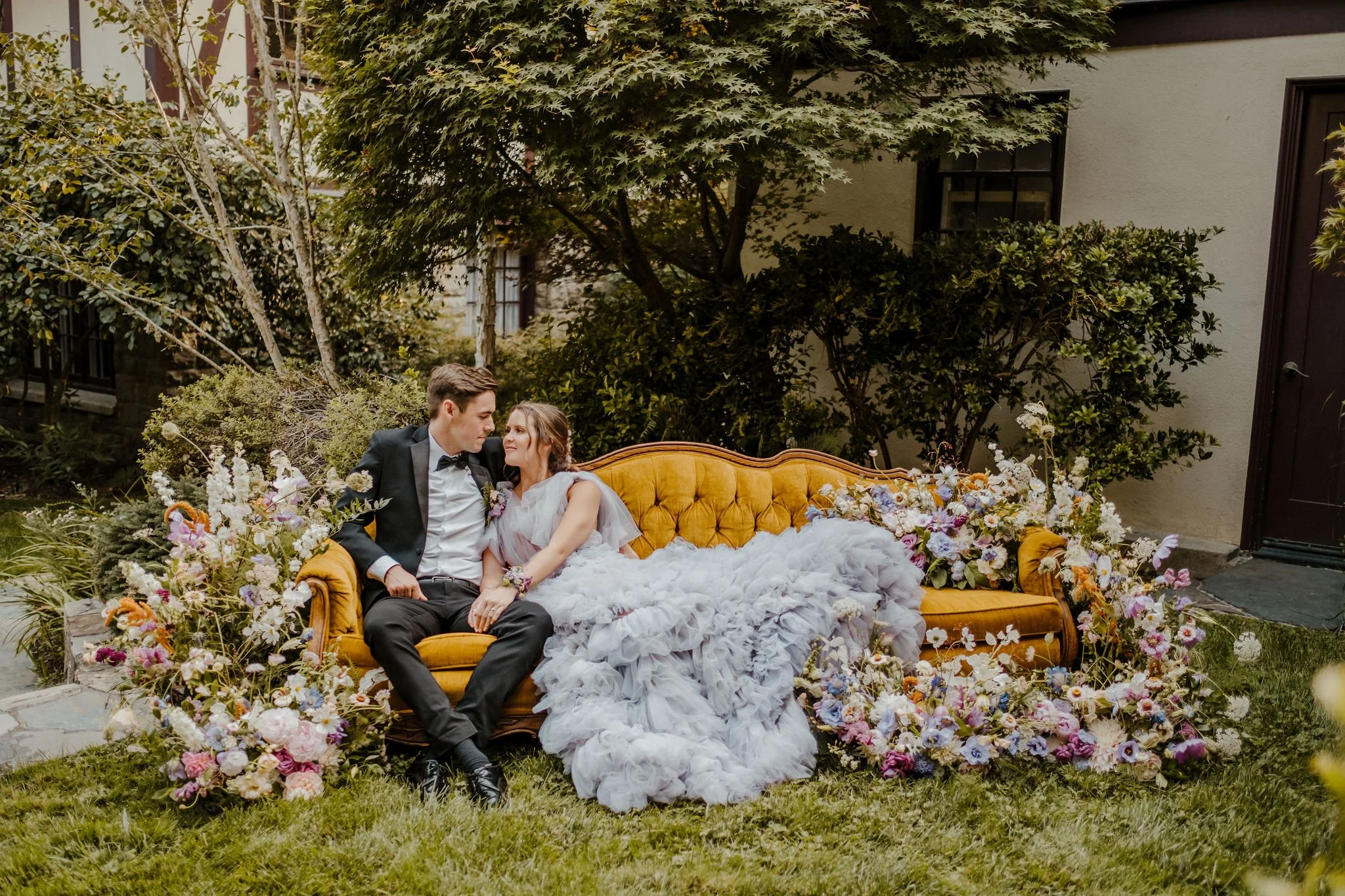 A bride and groom sitting on a vintage beige sofa surrounded by colorful flowers outdoors, with trees and the Julia Morgan Redwood Grove Great Room in the background.