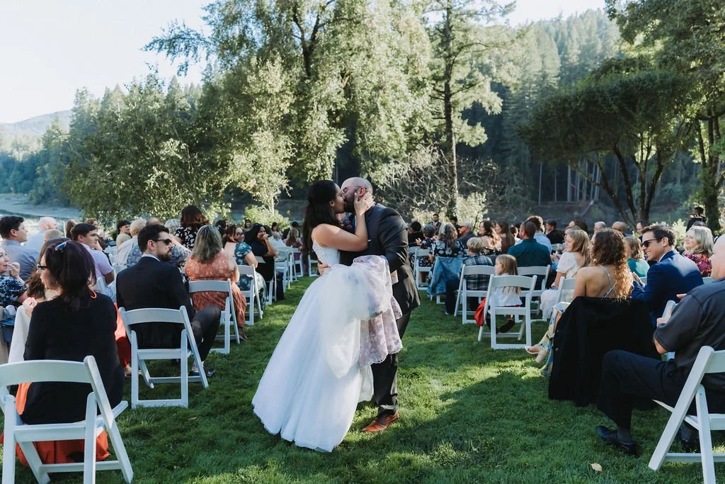 A wedding ceremony outdoors at the Julia Morgan Redwood Grove with a bride and groom kissing in the center, surrounded by seated guests on a grassy area with trees and a river in the background.