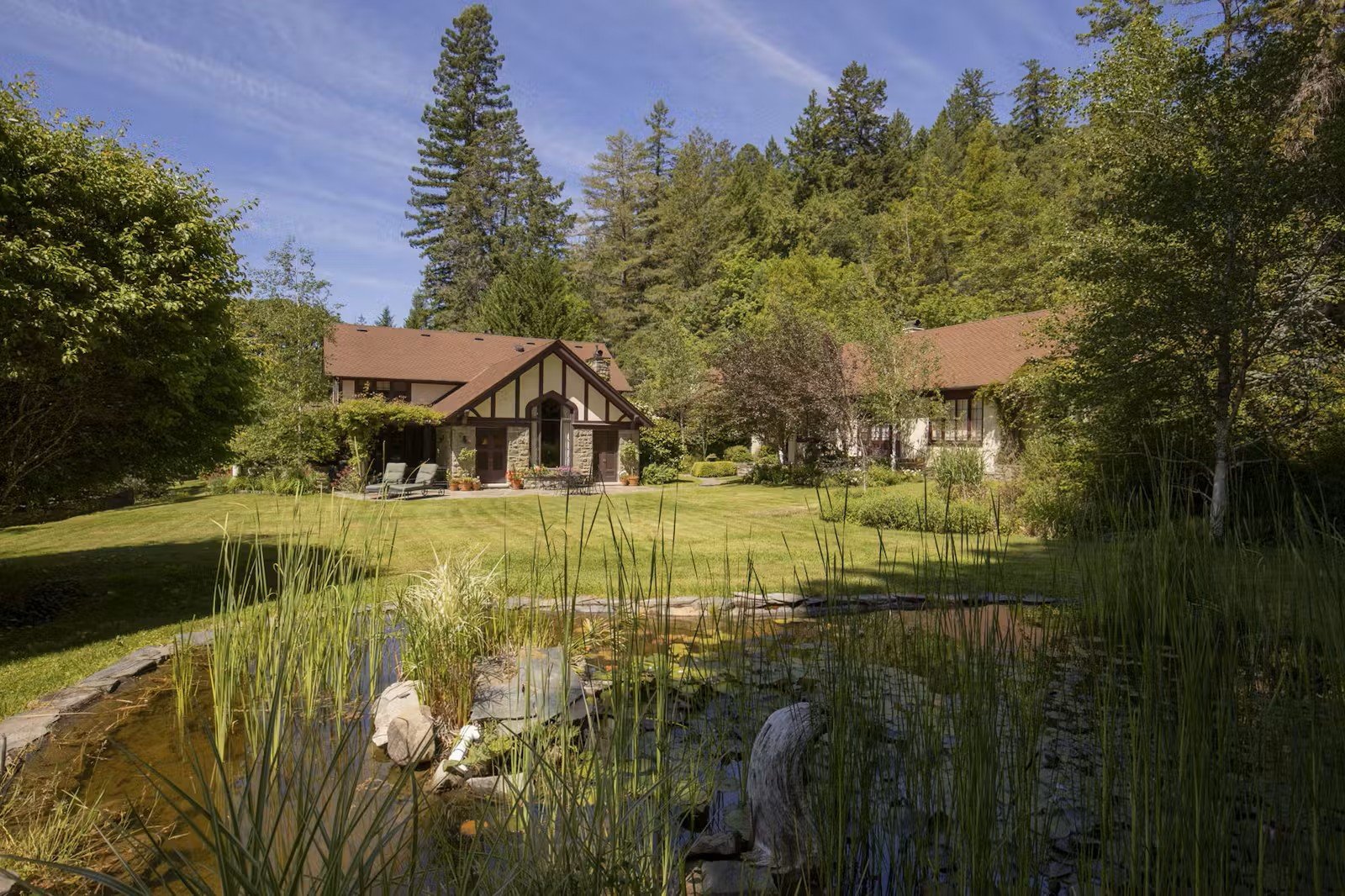 Large historic estate with a lush green lawn with a pond and tall trees under a partly cloudy sky.