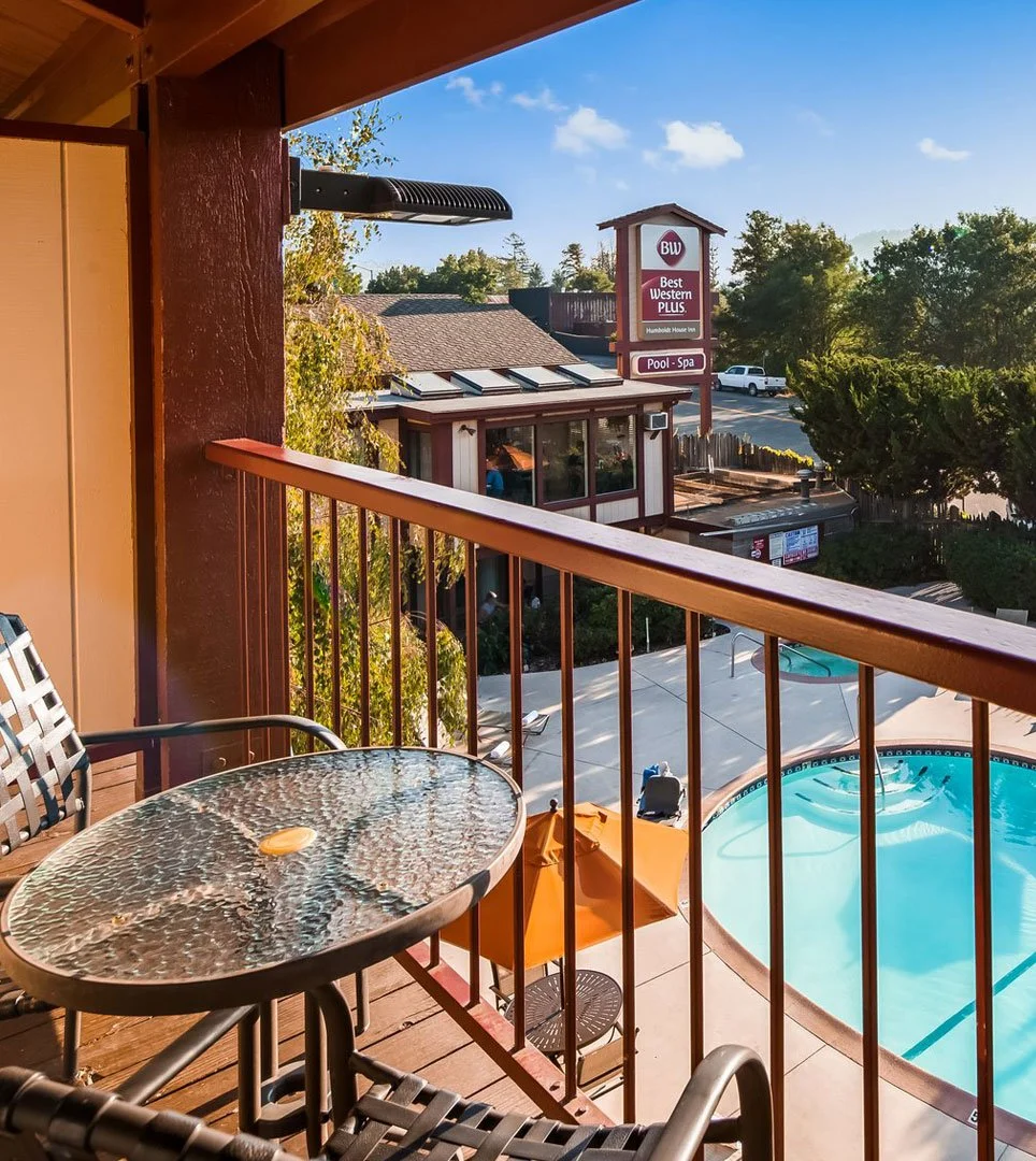 View from a balcony showing a swimming pool, neighboring building with a Best Western Plus sign, and trees under a blue sky with clouds.