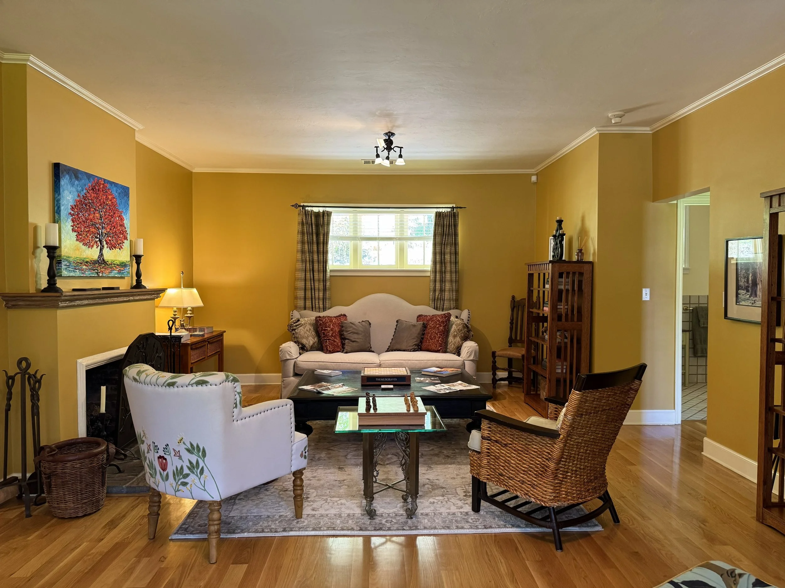 Living room with yellow walls, a white couch with pillows, a wooden coffee table with chess, a white armchair with floral patterns, a ceiling light fixture, a window with curtains, and artwork on the walls.