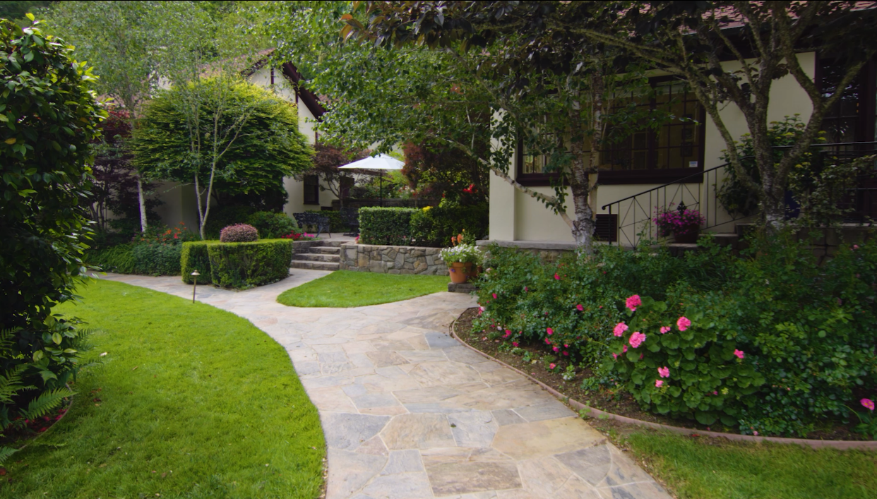 A well-maintained garden with a stone pathway, green grass, various trees and flowering bushes, and a house in the background with a patio and umbrella.