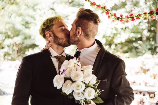 Two men in suits kissing, one holding a bouquet of white roses, outdoors with trees and decorative red flowers in the background.