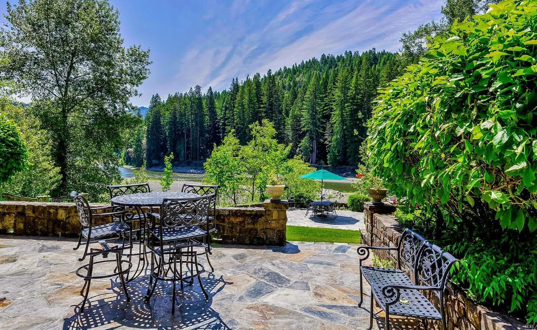 A stone terrace with metal patio furniture overlooks a lush green landscape with trees, the Eel river, and forested Humboldt County hills in the distance under a partly cloudy sky in the Benbow Valley.