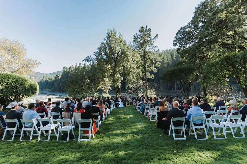 A large group of people attending an outdoor wedding ceremony at Julia Morgan Redwood Grove in a lush, green park surrounded by trees and a body of water in the background.