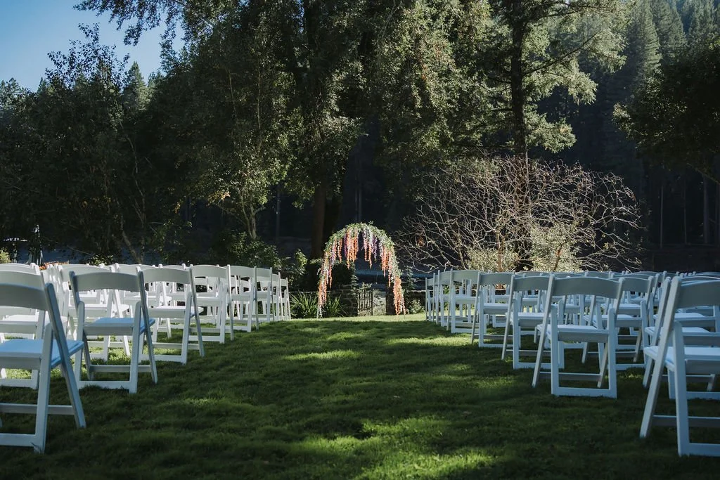 An outdoor wedding setup with white chairs arranged on a grassy lawn facing a floral arch at the altar, surrounded by trees.
