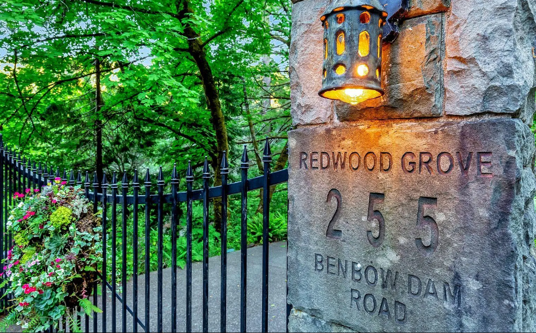 Stone entrance pillar with a lantern and engraved text reading 'Redwood Grove 255 Benbow Dam Road', next to a black iron fence with a flower basket, surrounded by lush green trees.