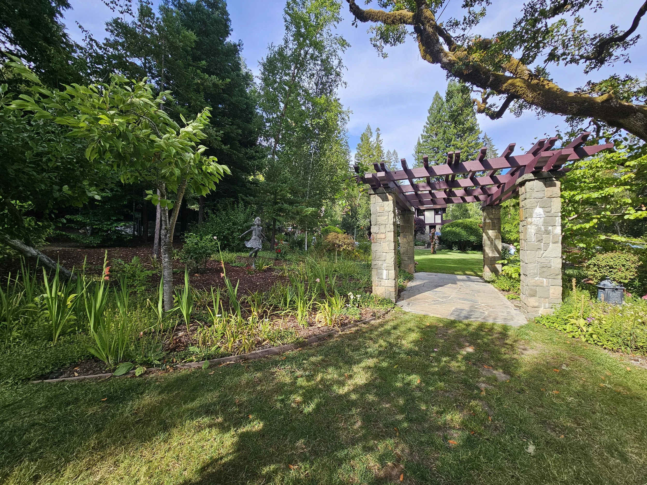 A garden with green trees, shrubs, plants, and a stone archway with a purple wooden trellis on top, under a blue sky with some clouds.
