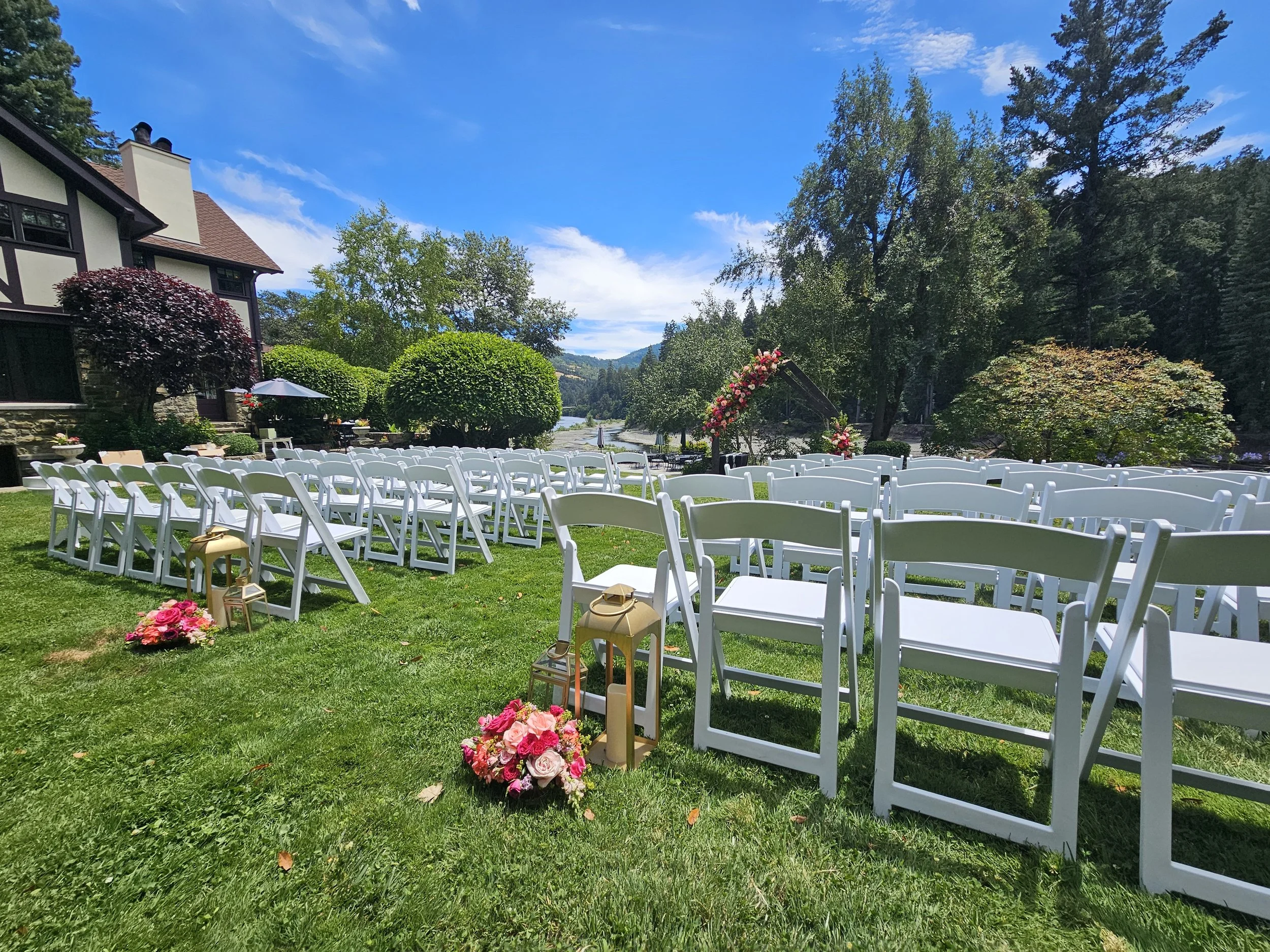 An outdoor wedding setup with white chairs arranged on a grassy lawn, pink floral arrangements, lanterns, and a decorative arch overlooking a river, surrounded by trees and mountains under a clear blue sky.